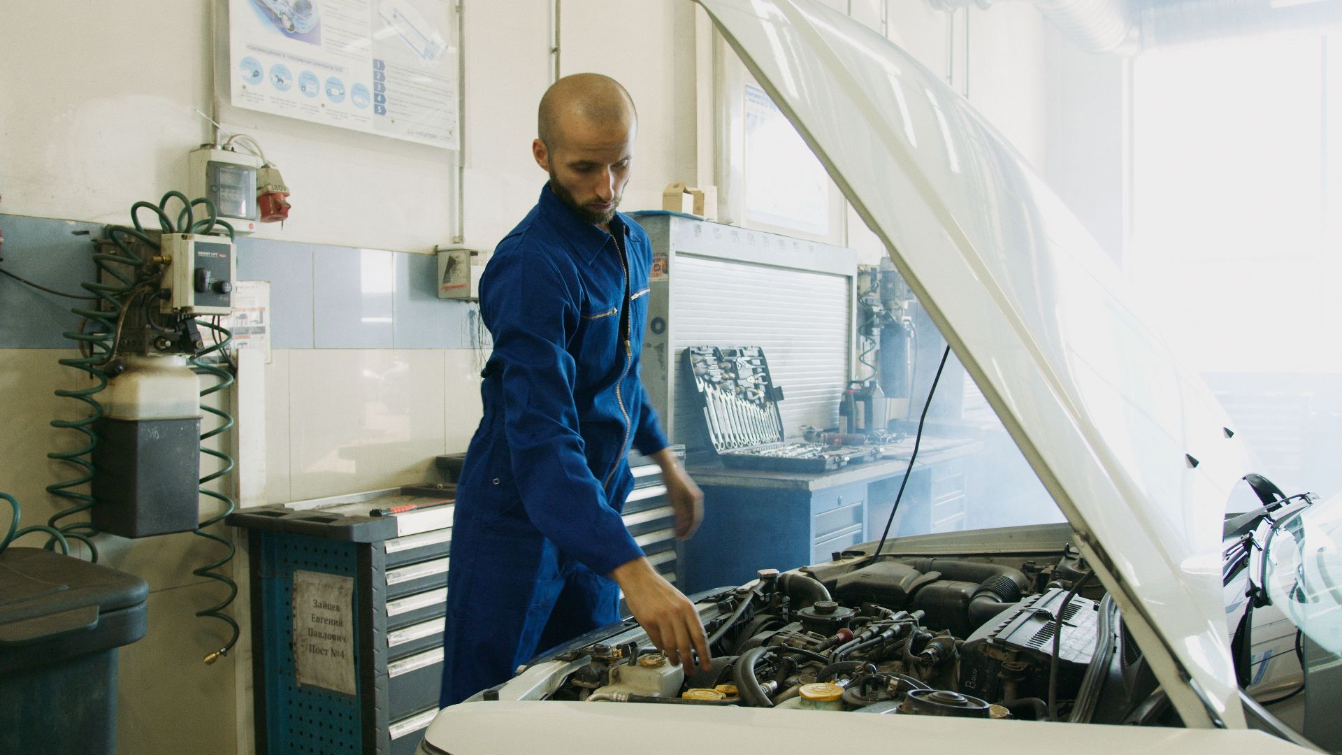 Mechanic in blue uniform inspecting a car engine in a workshop.