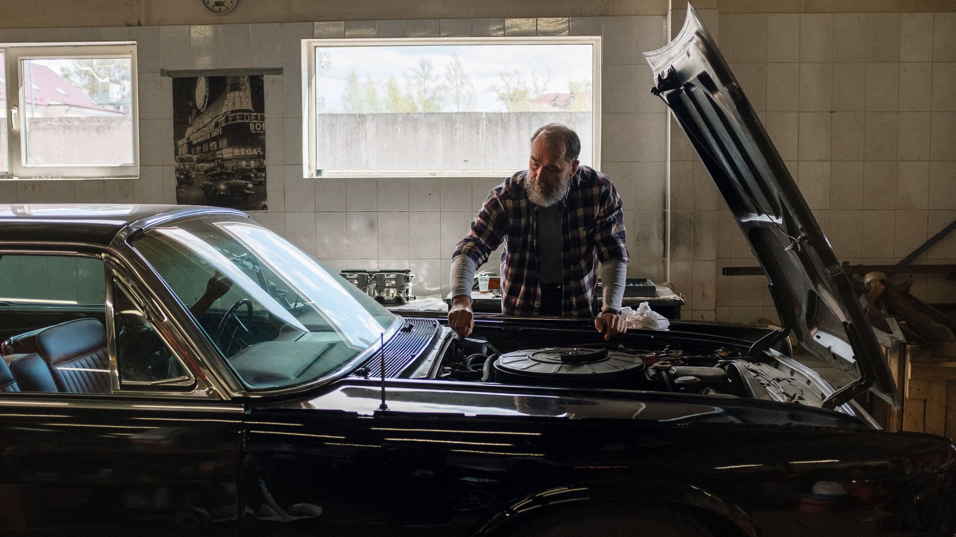 A bearded mechanic examines a classic car engine in a well-lit auto repair shop.