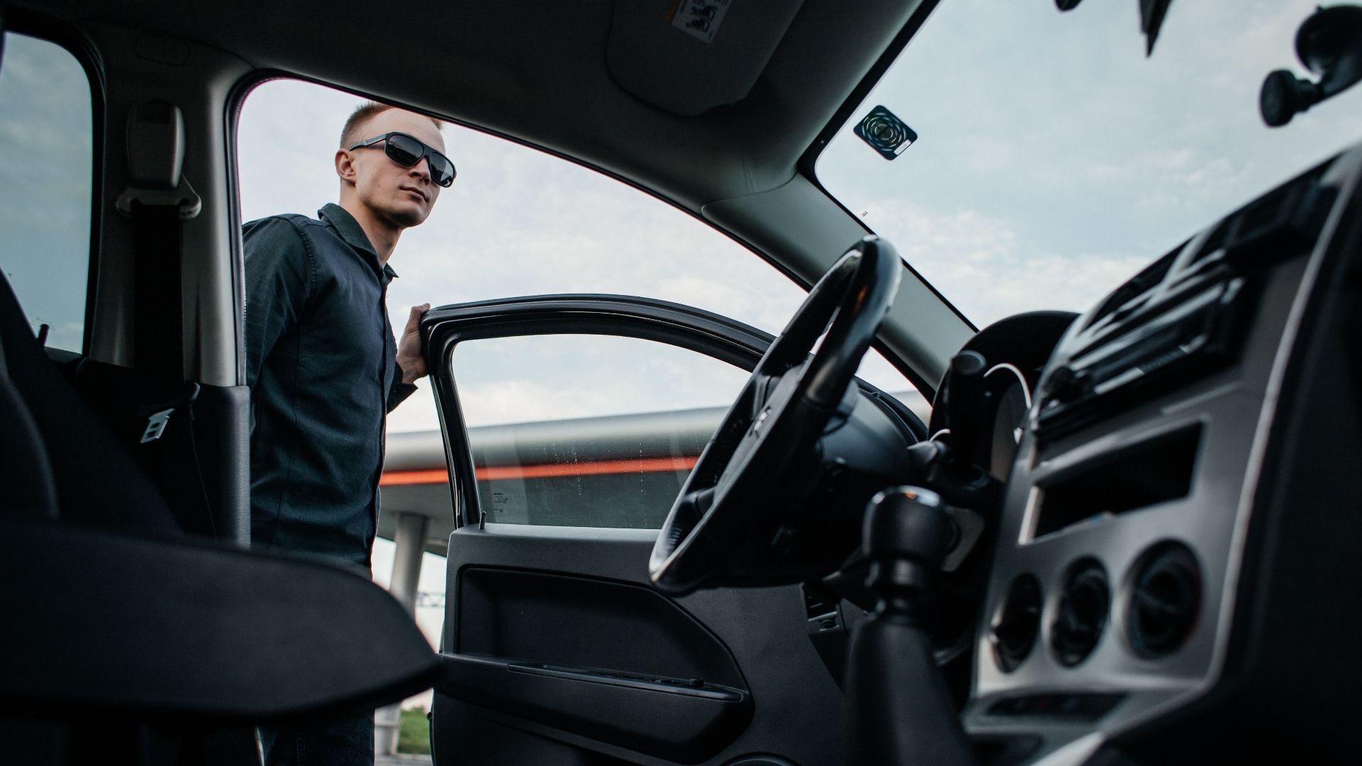 A man in sunglasses stands by an open car door, viewed from the interior.