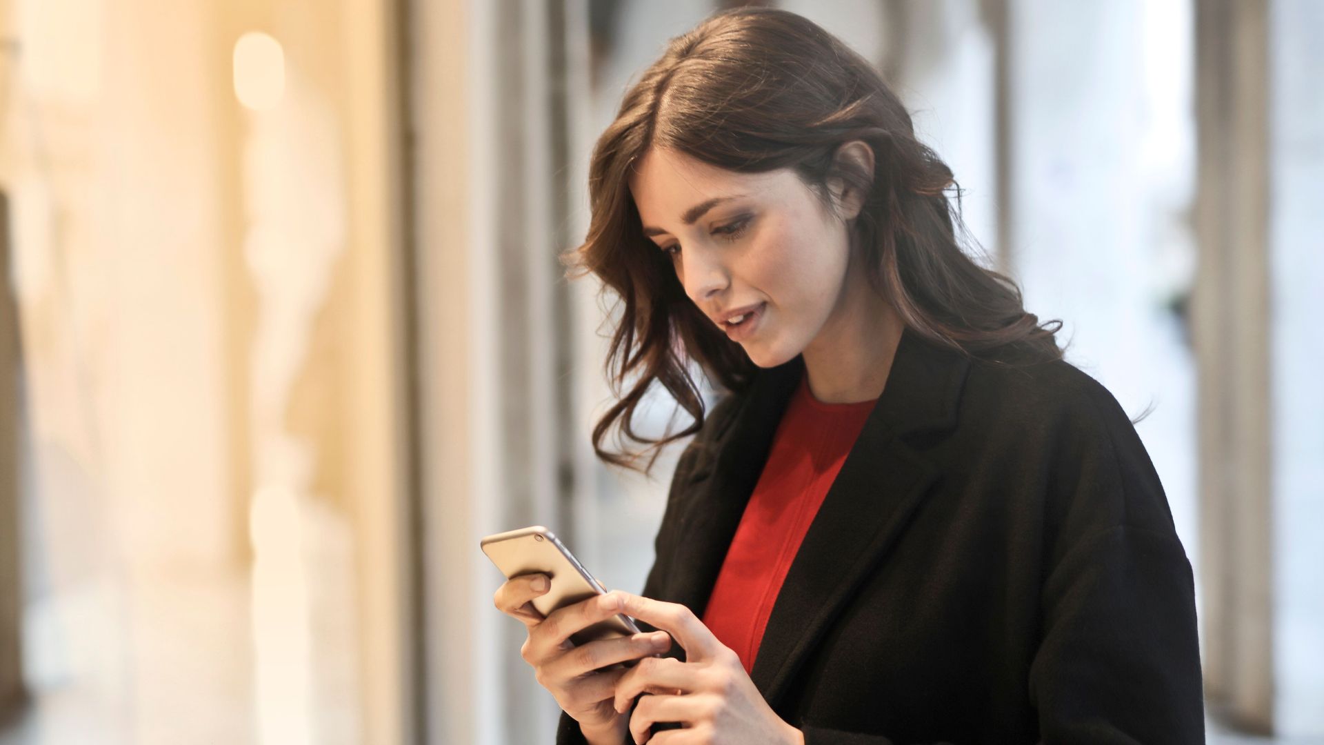 Young woman using smartphone at a store window reflecting her image, in casual fashion attire.