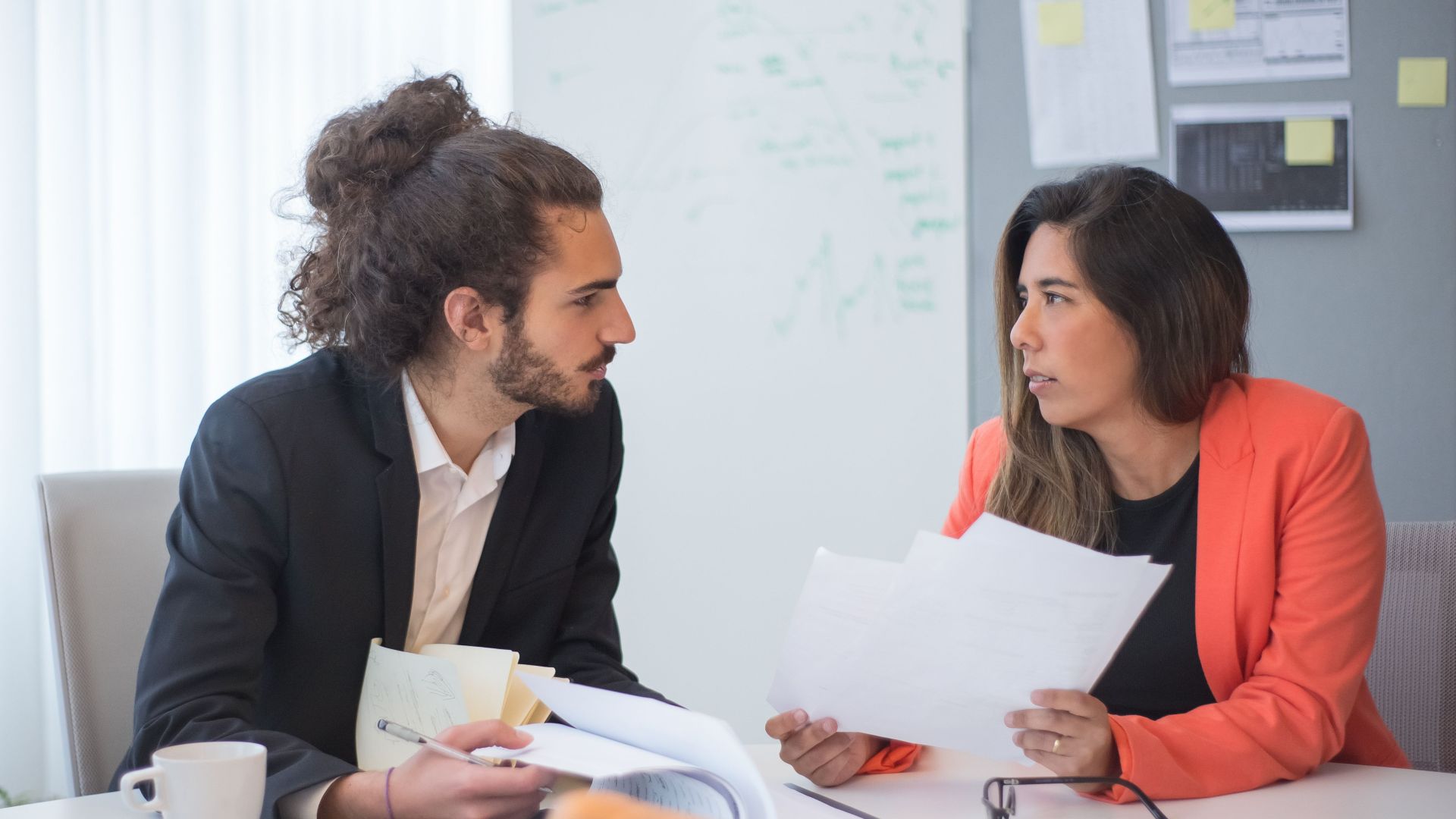 Two colleagues engaged in a discussion at an office table with documents and coffee.