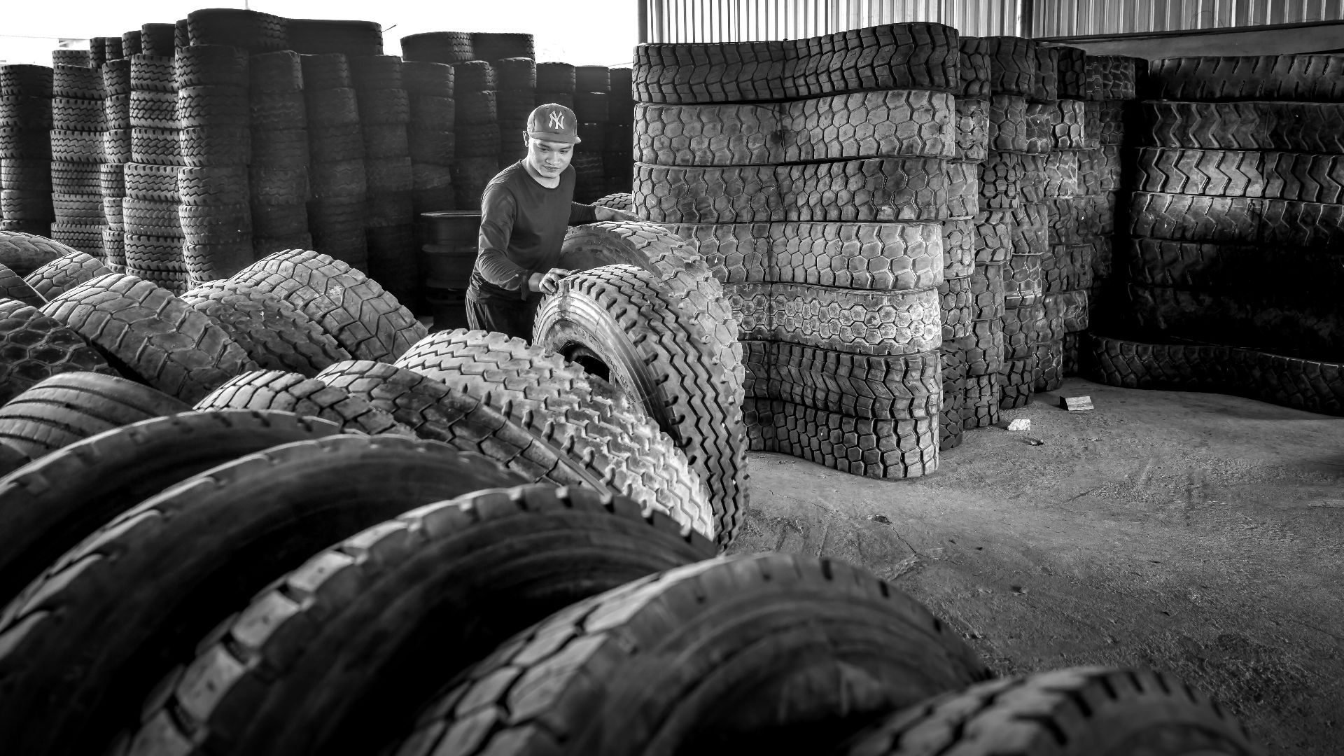 An Asian man working in a monochrome warehouse filled with stacked tires, emphasizing industrial labor.