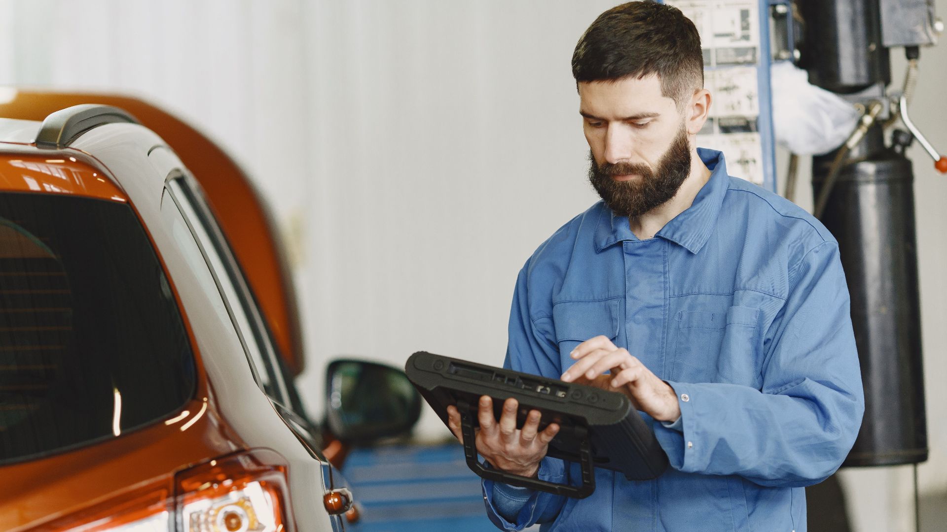 A mechanic in coveralls using a diagnostic tool at an auto repair shop beside a car.