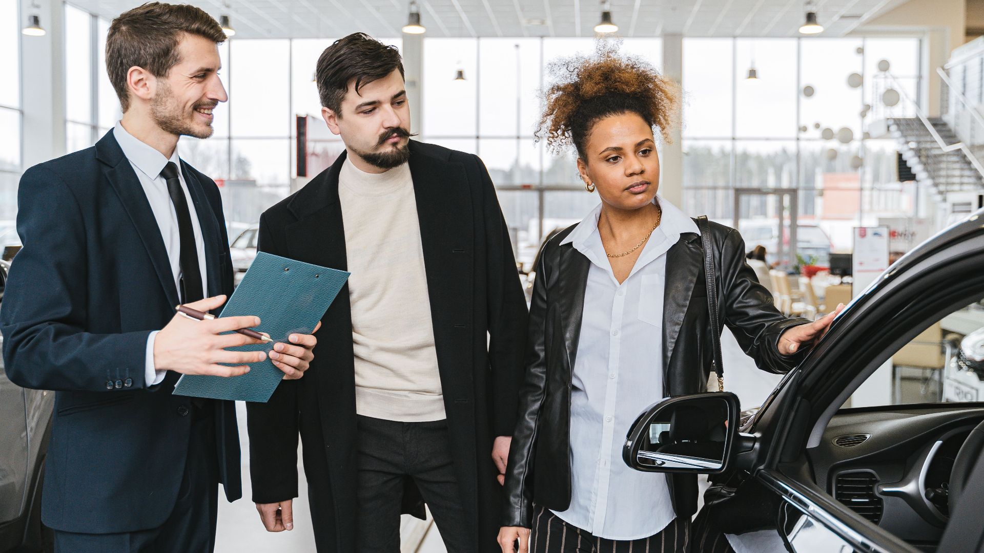 An interracial couple consults with a salesman at a car dealership, exploring vehicle options.