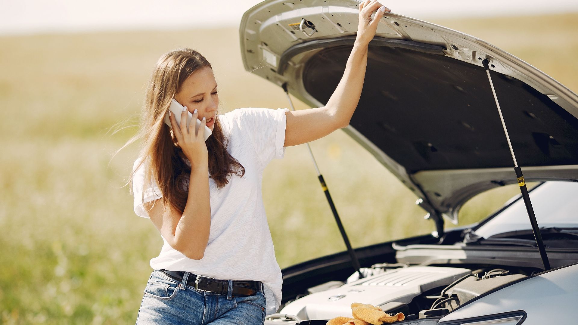 Worried young female driver in white t shirt and jeans speaking on phone while leaning on broken car with open hood asking for help with repair against green field in sunny summer day