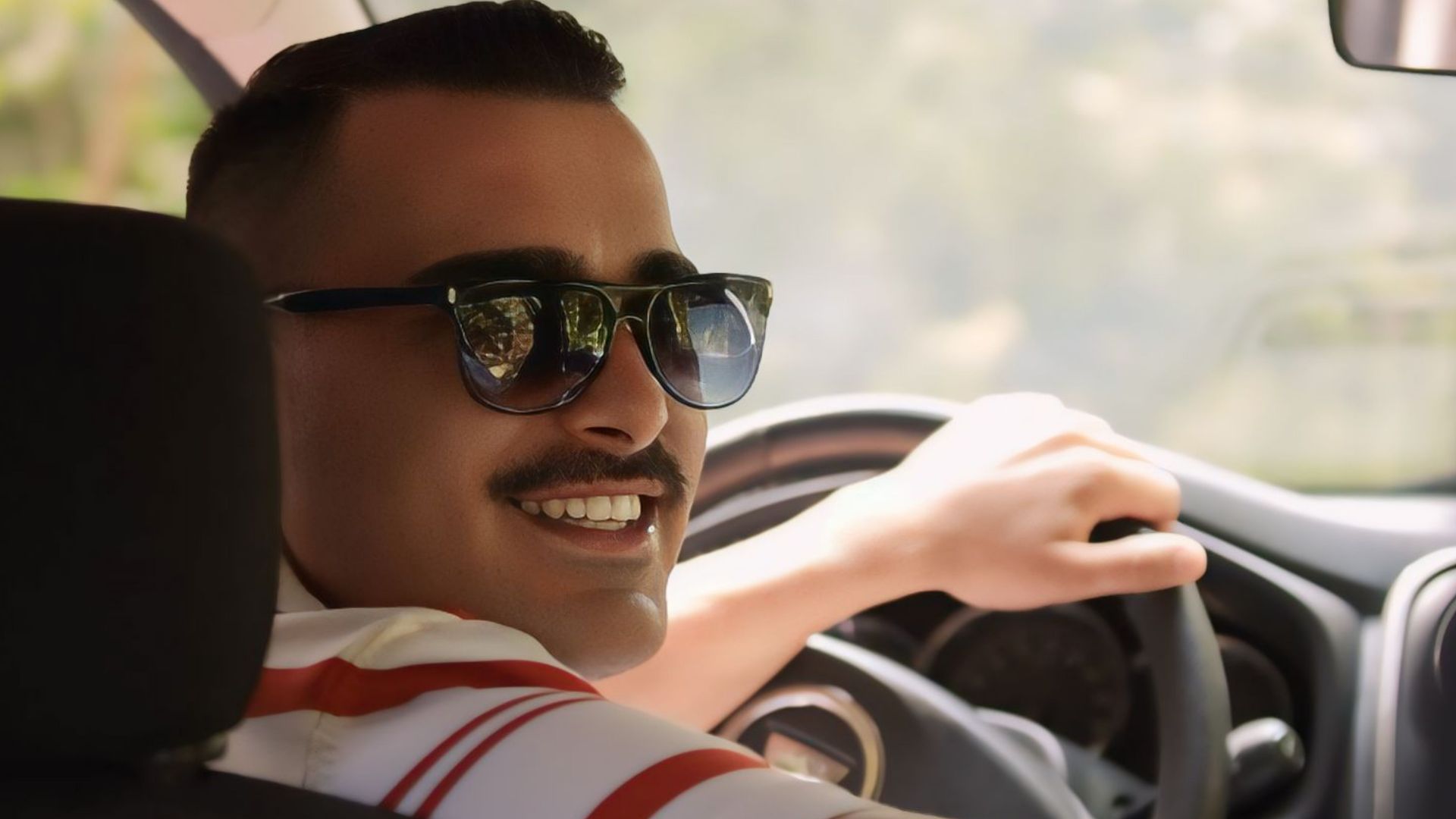Smiling man wearing sunglasses driving a car with sunlight streaming through the window.