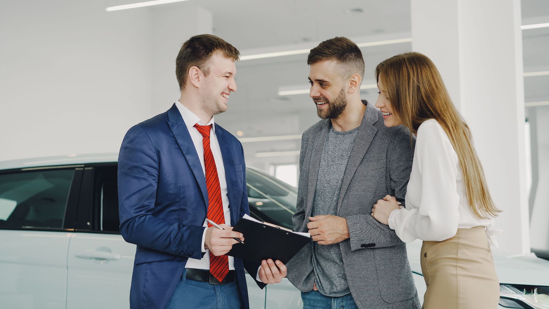 A smiling couple buys a new car from a confident salesman inside a modern car dealership.
