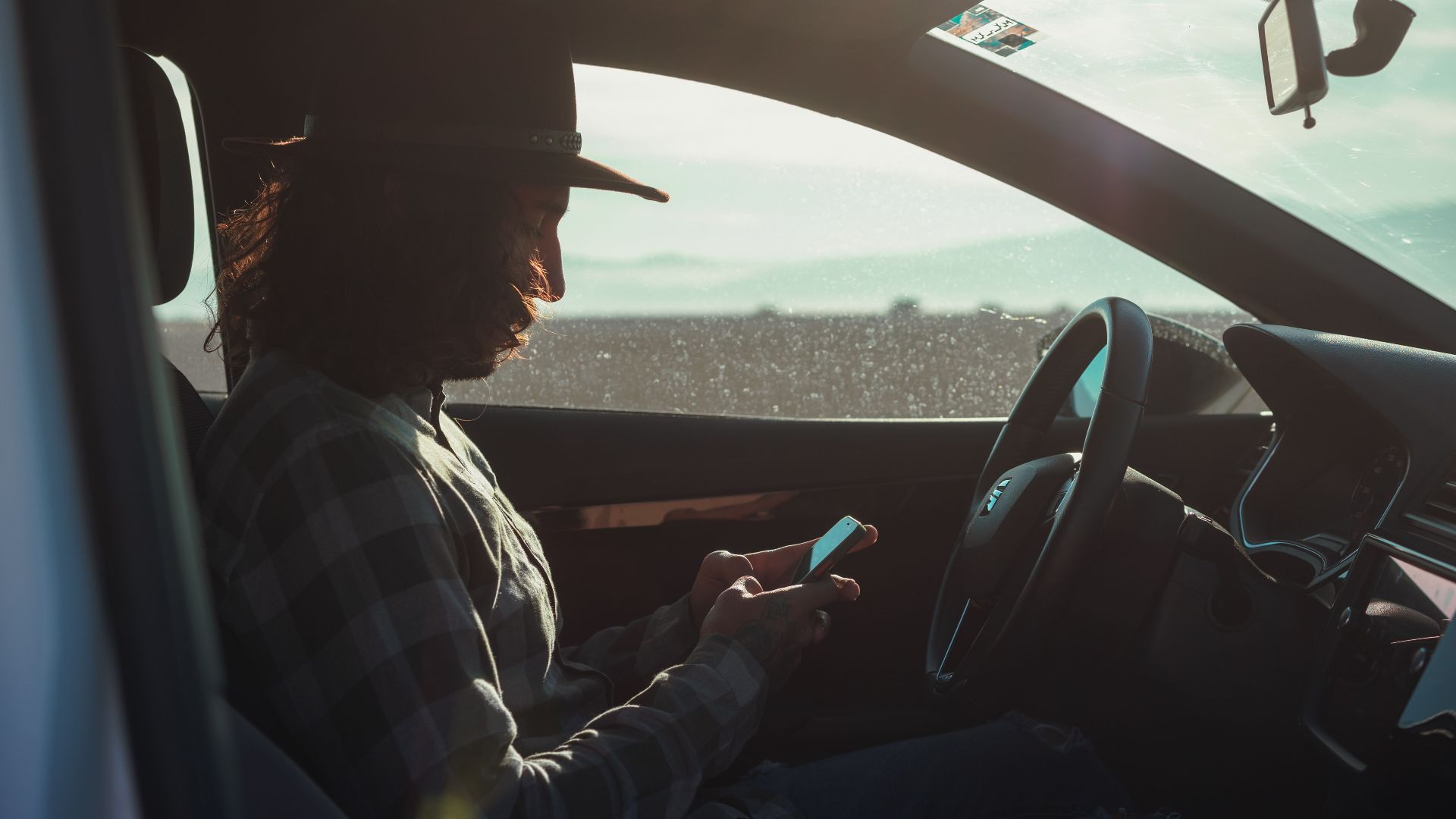 Profile of a man with a hat using a phone inside a car during the day.