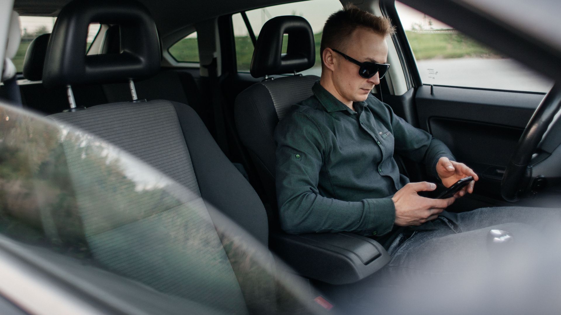 Young man using smartphone inside a parked car, wearing sunglasses.