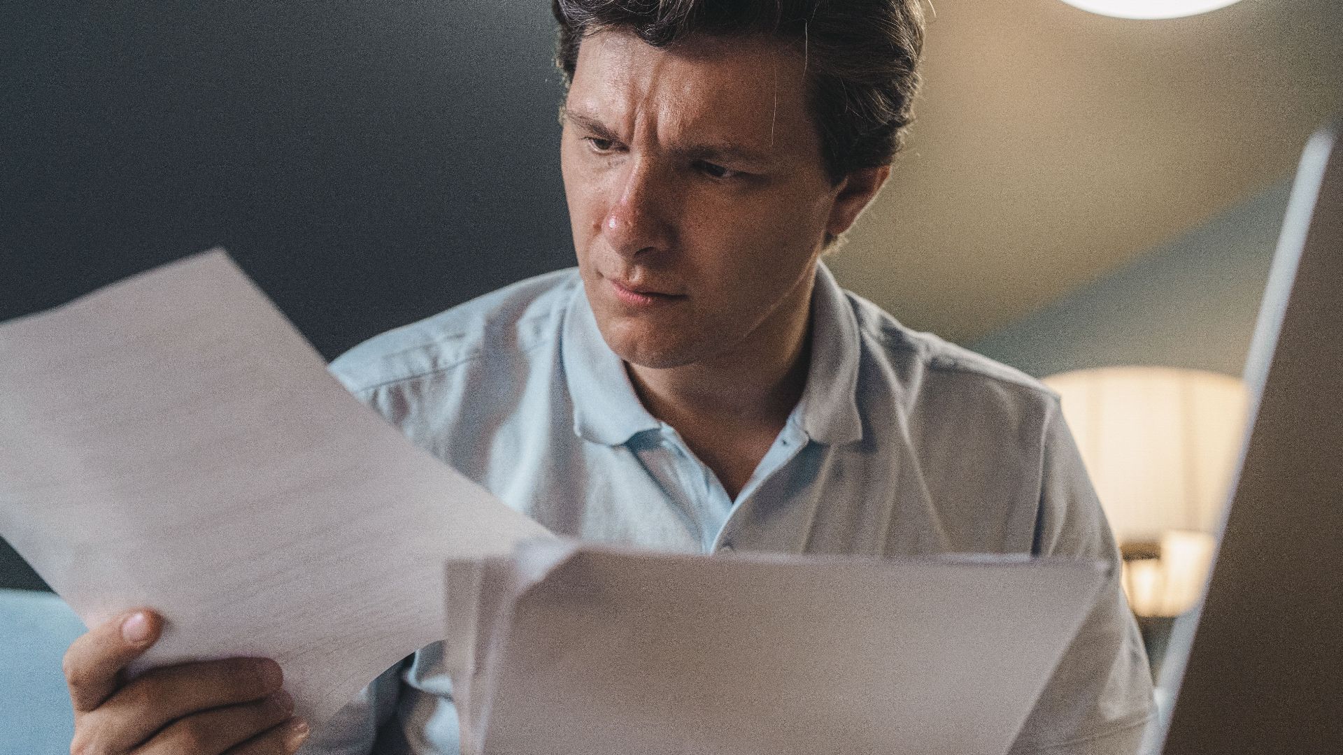A man intently examines papers, seated indoors under warm lighting, focusing on his work.