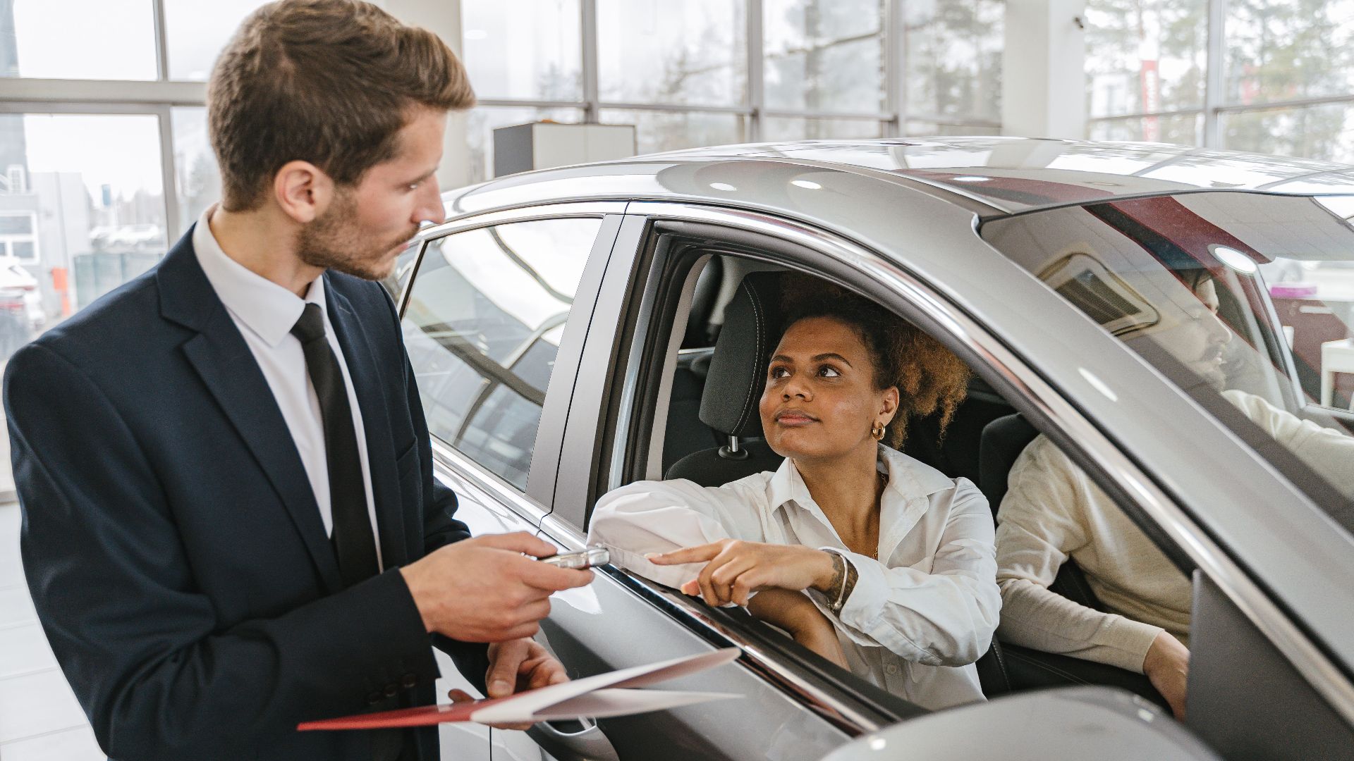 A car dealer hands keys to a woman sitting inside a vehicle in a car showroom.