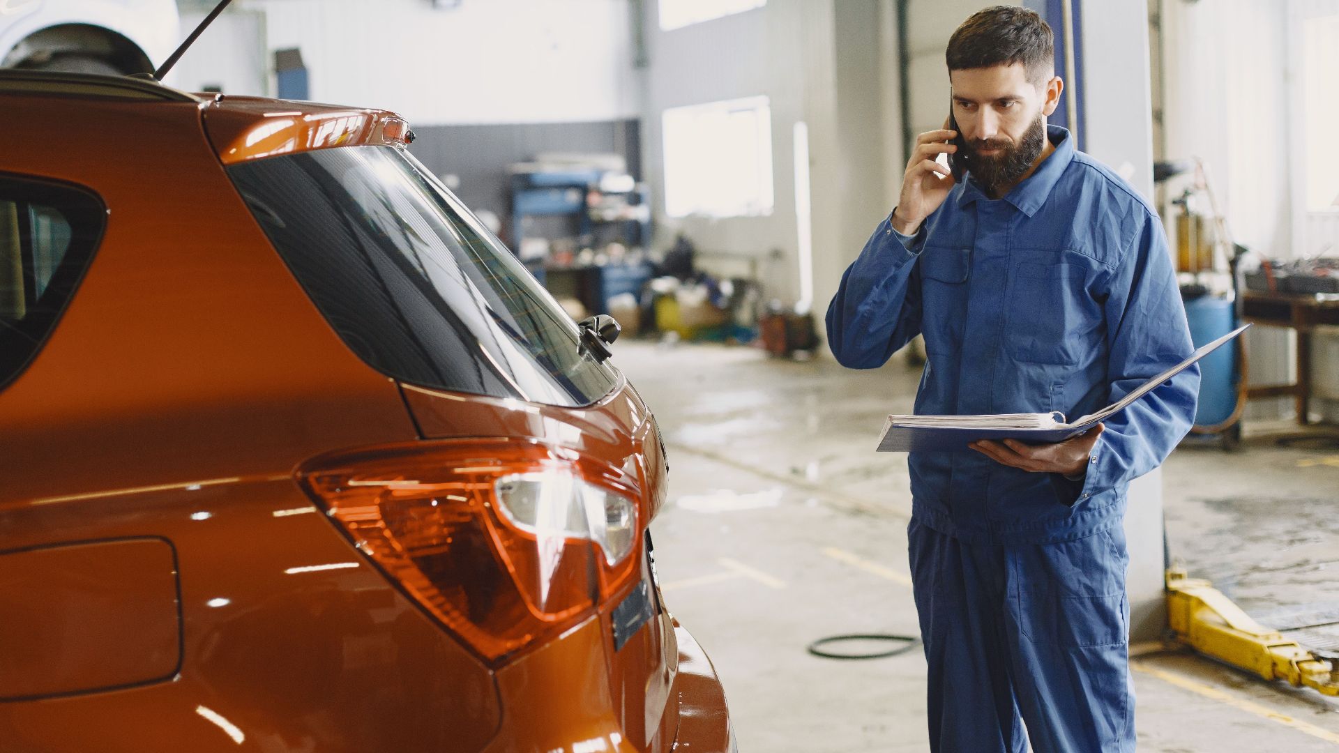 Mechanic in a blue uniform talks on phone beside a red car in an auto repair shop.