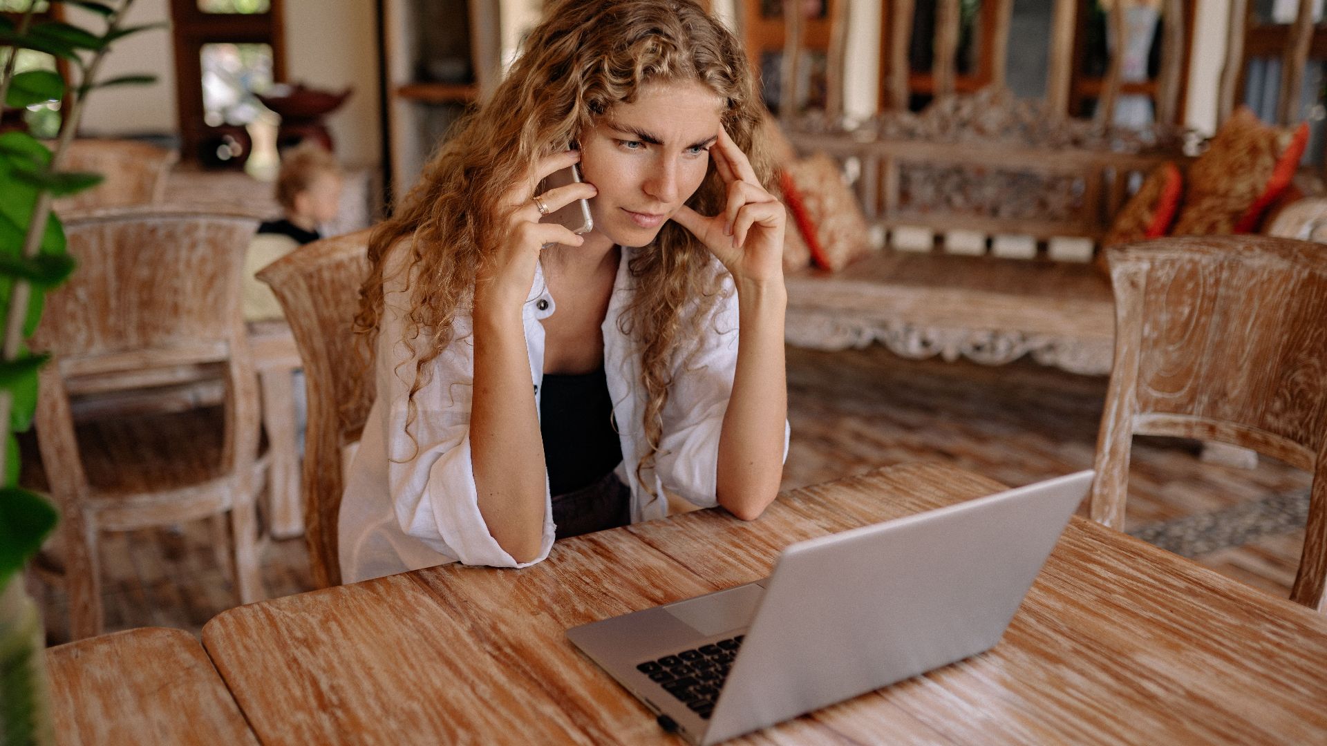 Focused woman multitasking with a phone and laptop in a cozy, rustic setting.