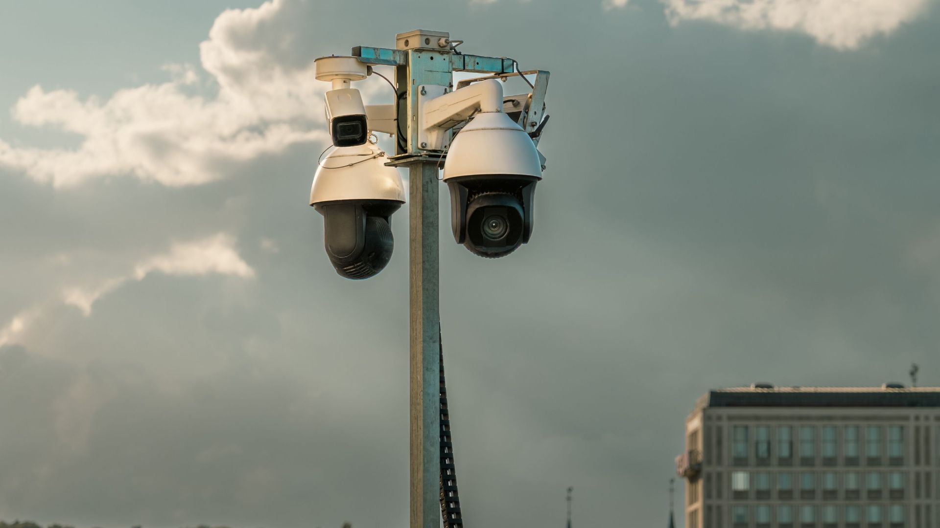 Two modern surveillance cameras mounted on a pole in an urban setting under a cloudy sky.