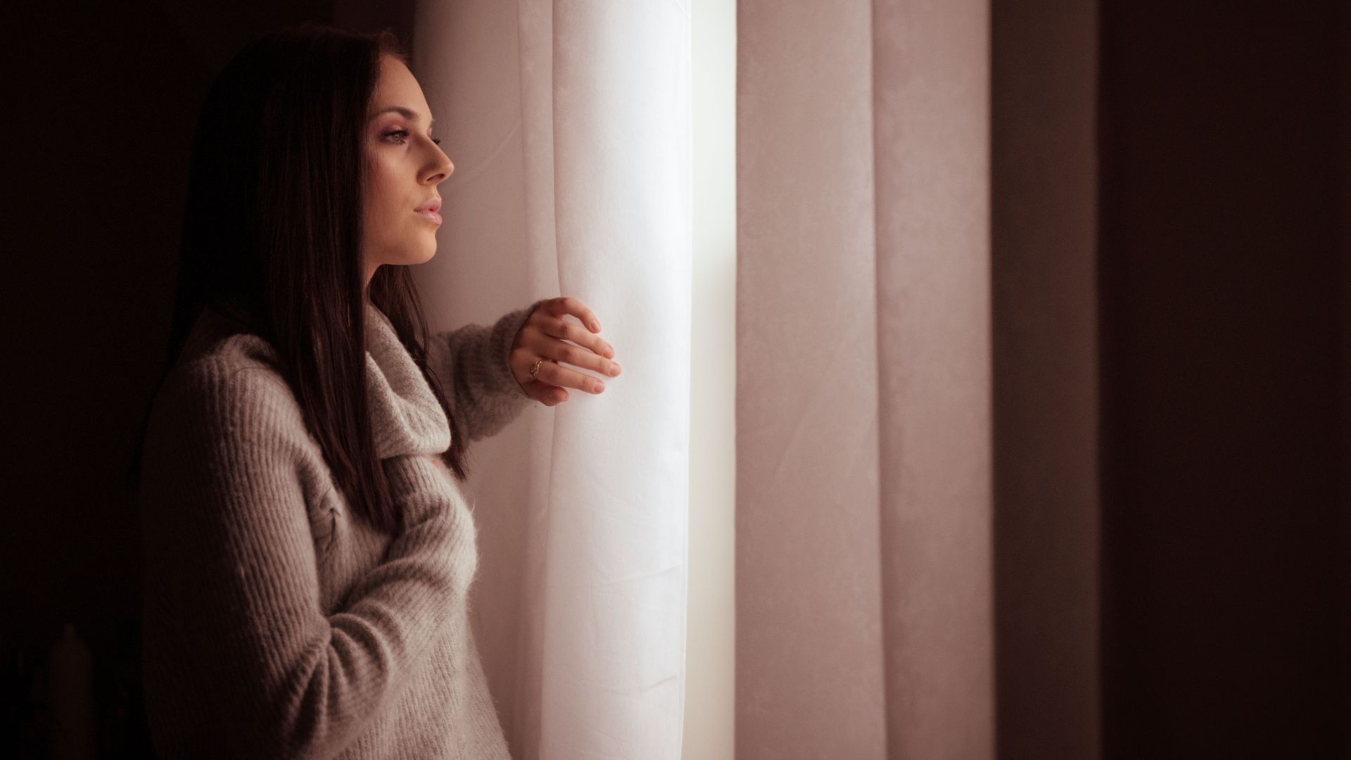 A woman in a sweater thoughtfully gazes out of a sunlit window, creating a warm indoor atmosphere.