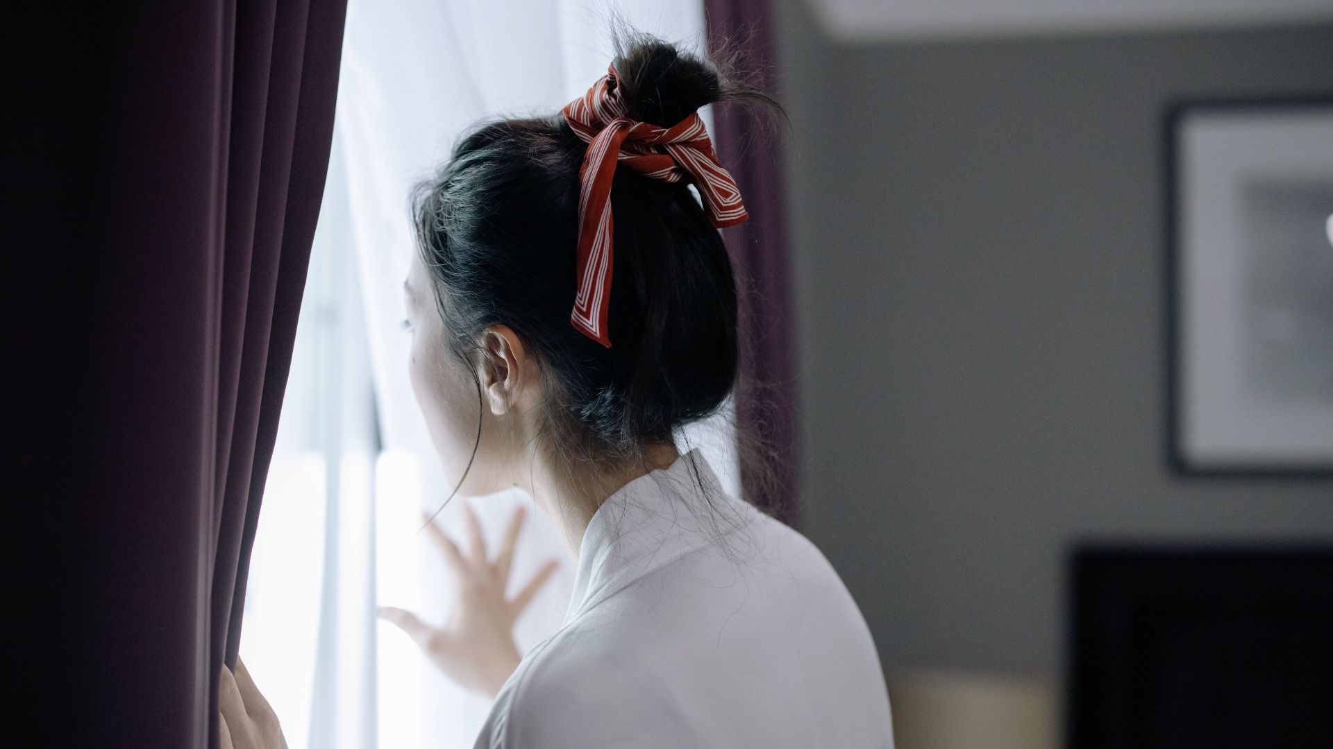 A woman with a hair bun looks outside through a window, creating a serene indoor scene.