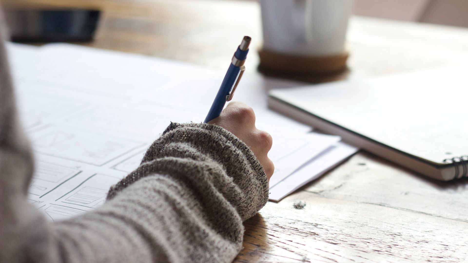 person writing on brown wooden table near white ceramic mug