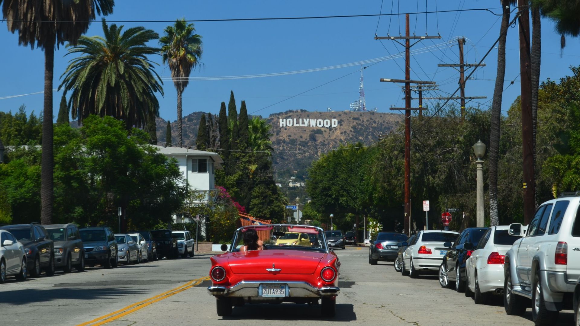 Red convertible driving down a sunny street towards the iconic Hollywood sign in Los Angeles.