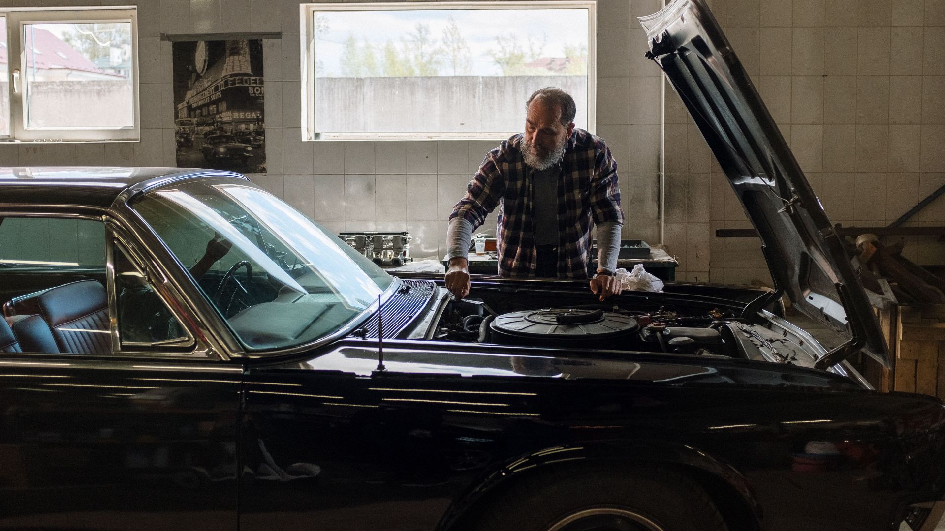 A bearded mechanic examines a classic car engine in a well-lit auto repair shop.