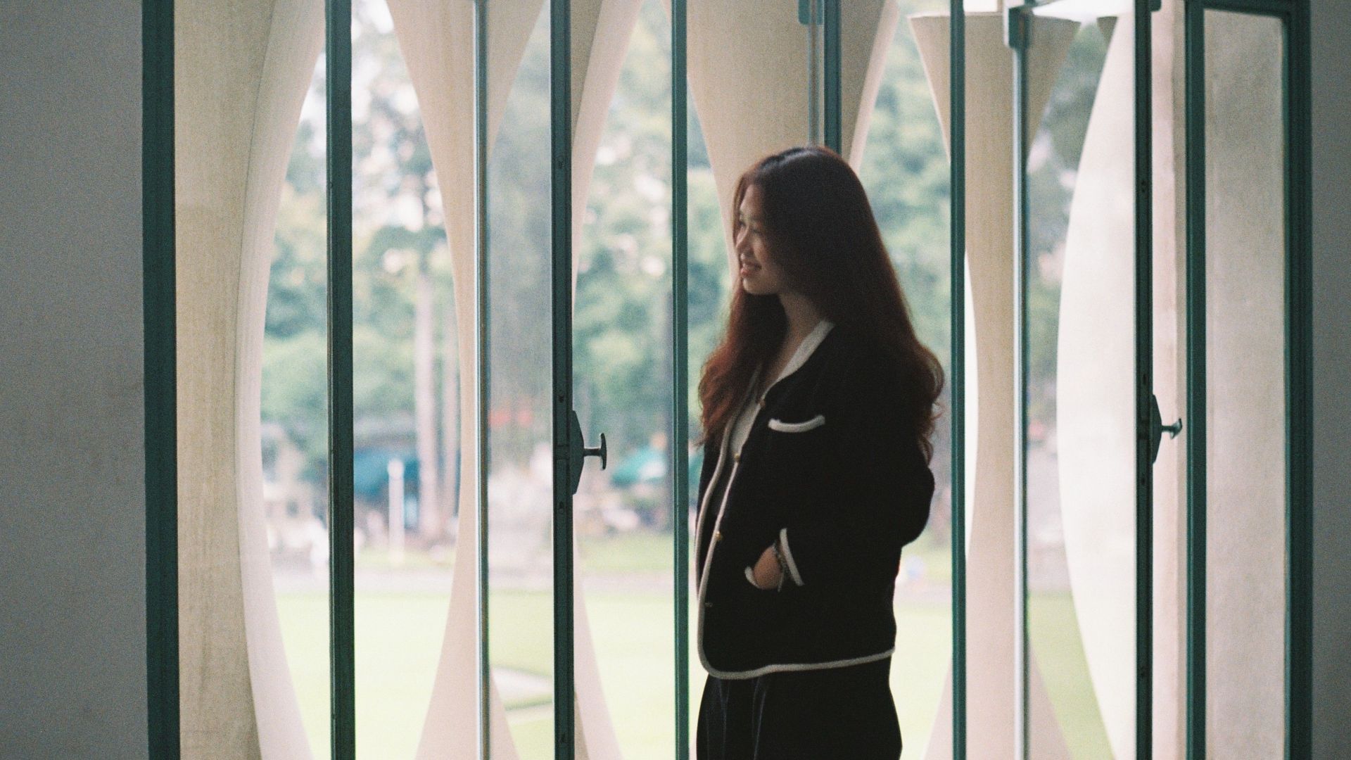 Fashionable Asian woman in black attire smiling by windows indoors during the day.