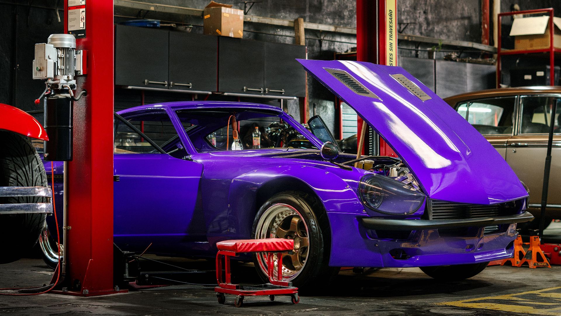 A classic car with a vibrant purple color in a mechanical workshop with its hood open.