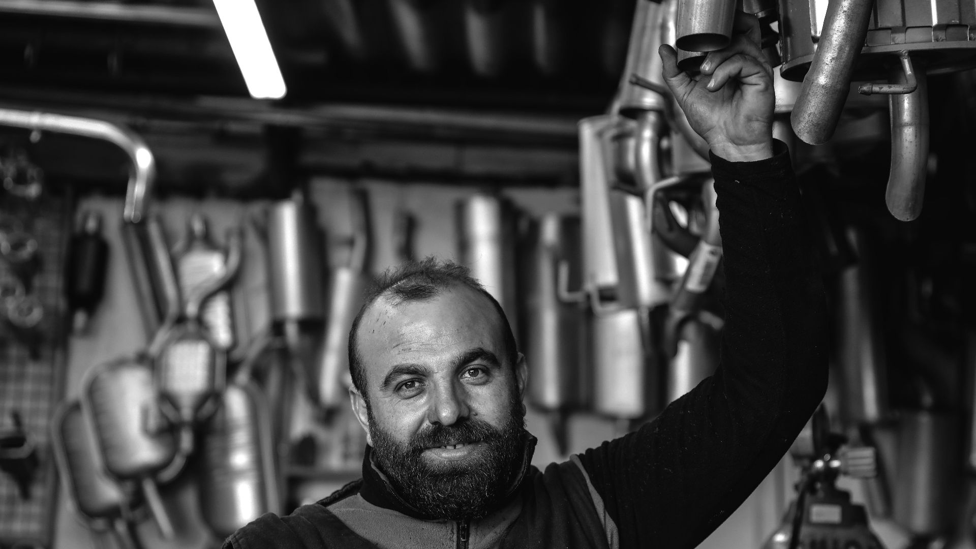 A smiling mechanic holding an exhaust pipe in a car repair garage. Black and white photo.