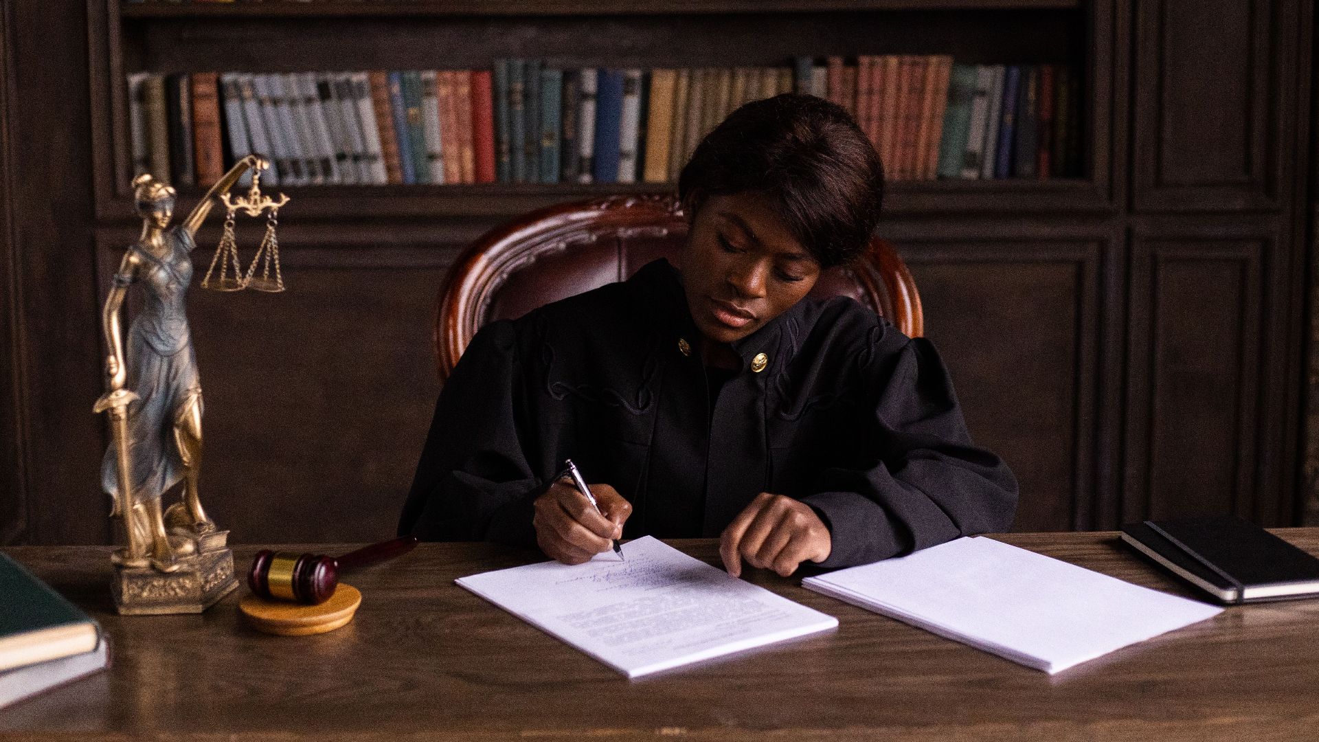 A judge in robes writing on a document at a desk in an office library with law books.