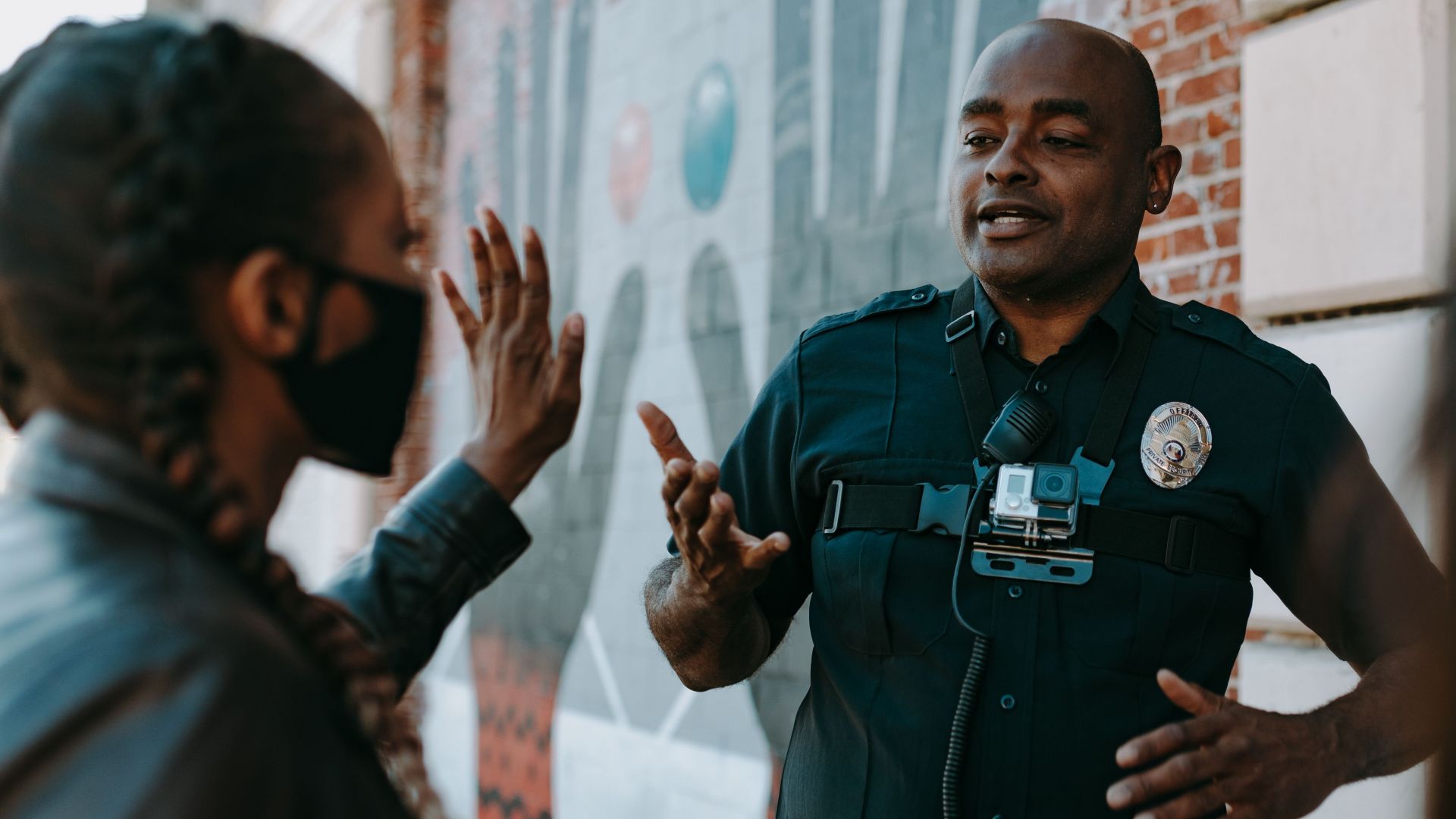 Police officer conversing with people on the street near a brick wall mural.