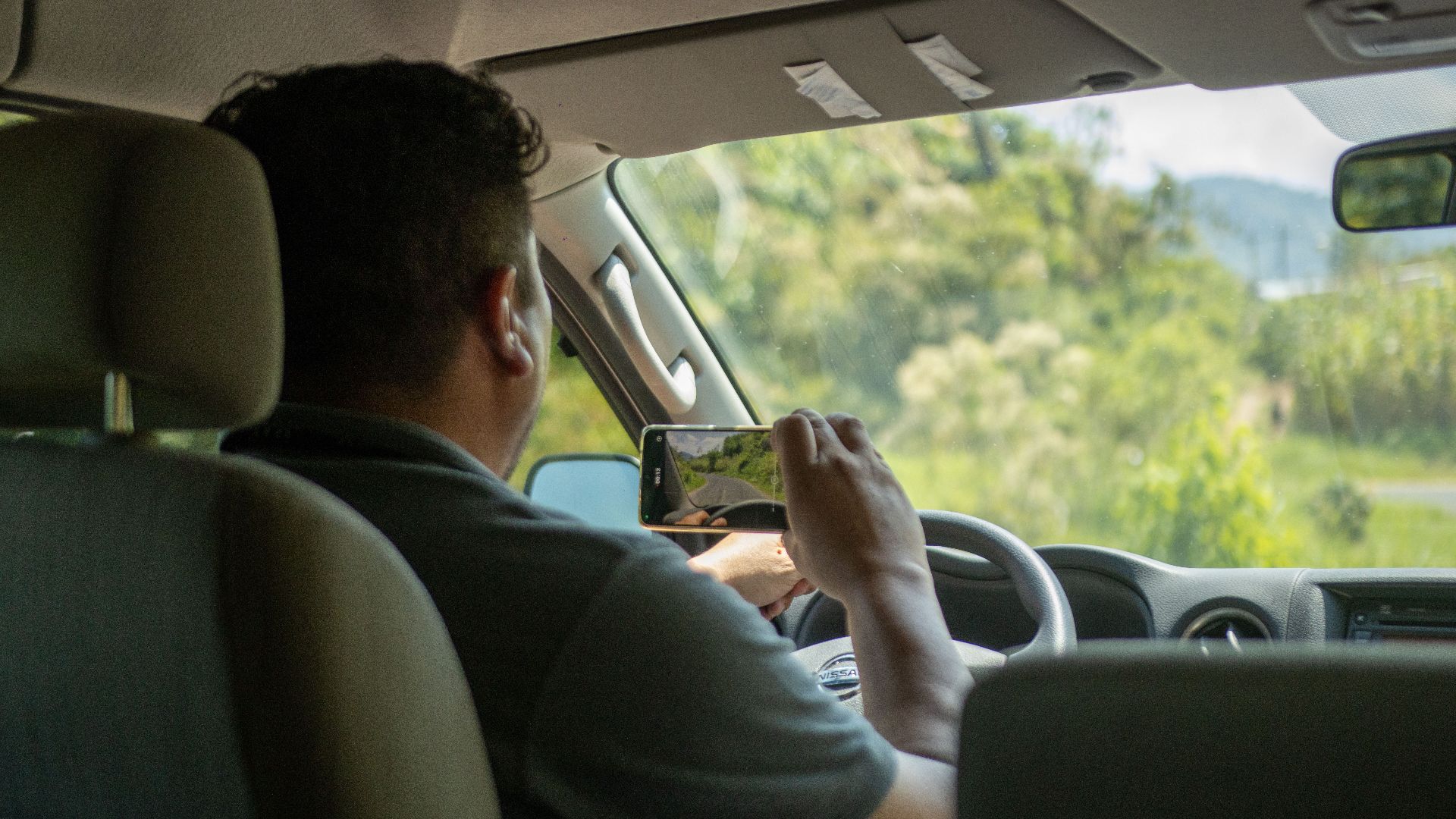 A man uses a smartphone to capture a scene outside while driving in a car interior during the day.
