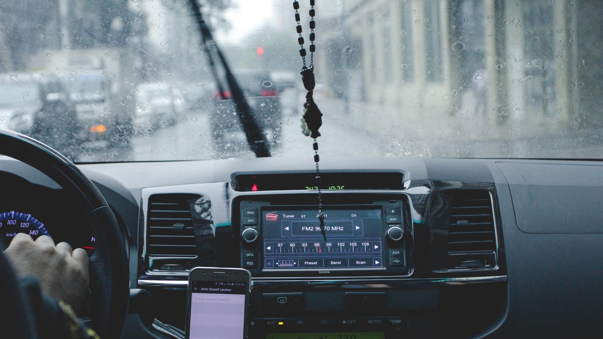 Car interior with driver navigating rainy cityscape using phone and dashboard.