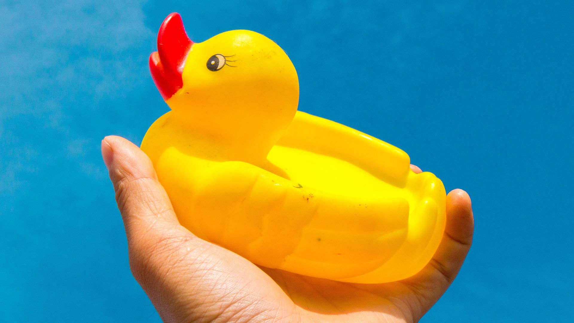 Hand holding a yellow rubber duck against a clear blue sky, symbolizing fun and playfulness.