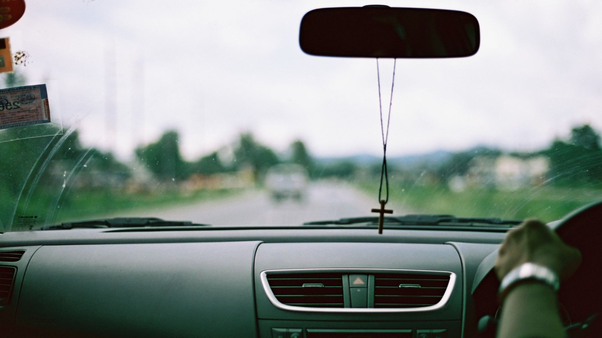 Interior view of a car dashboard with hanging cross on rear-view mirror, blurred road ahead.