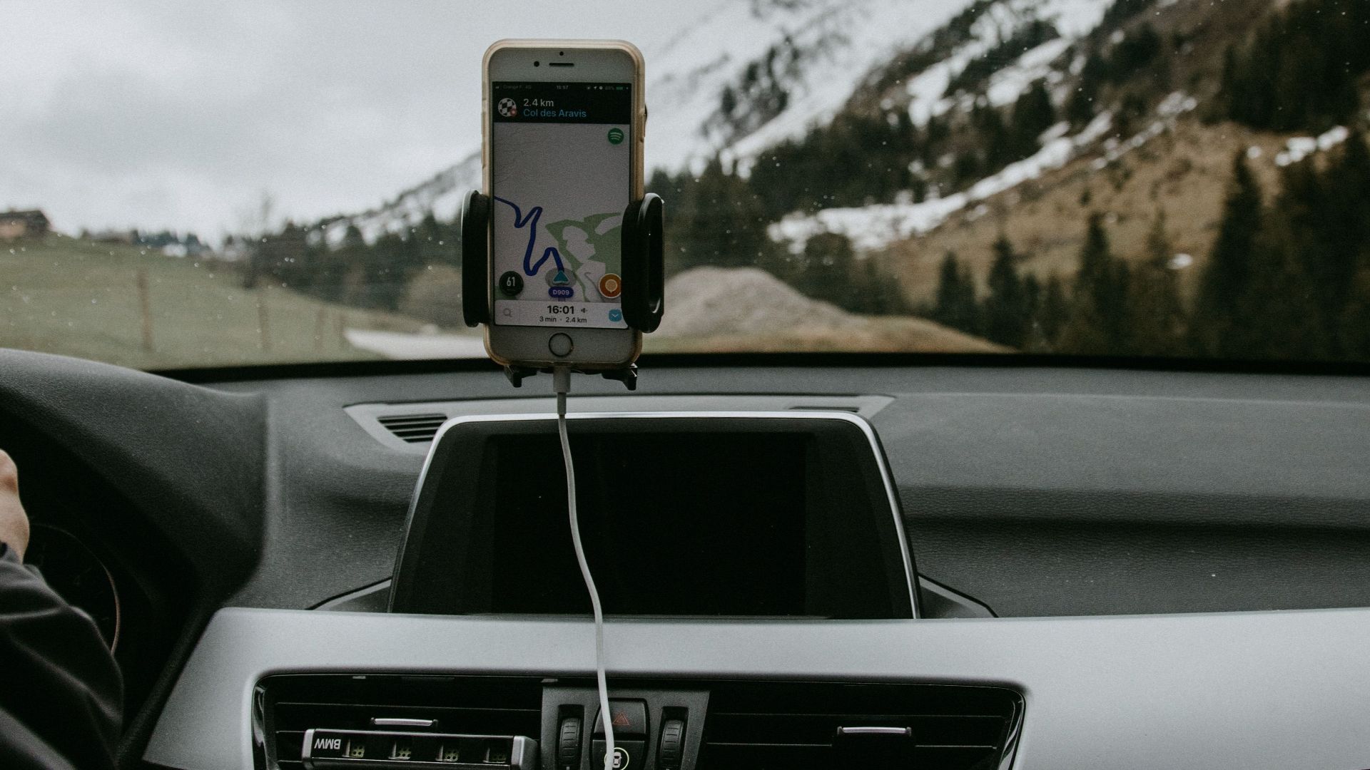 Driver in car with smartphone showing route due to GPS against snowy hill slope with coniferous trees and road
