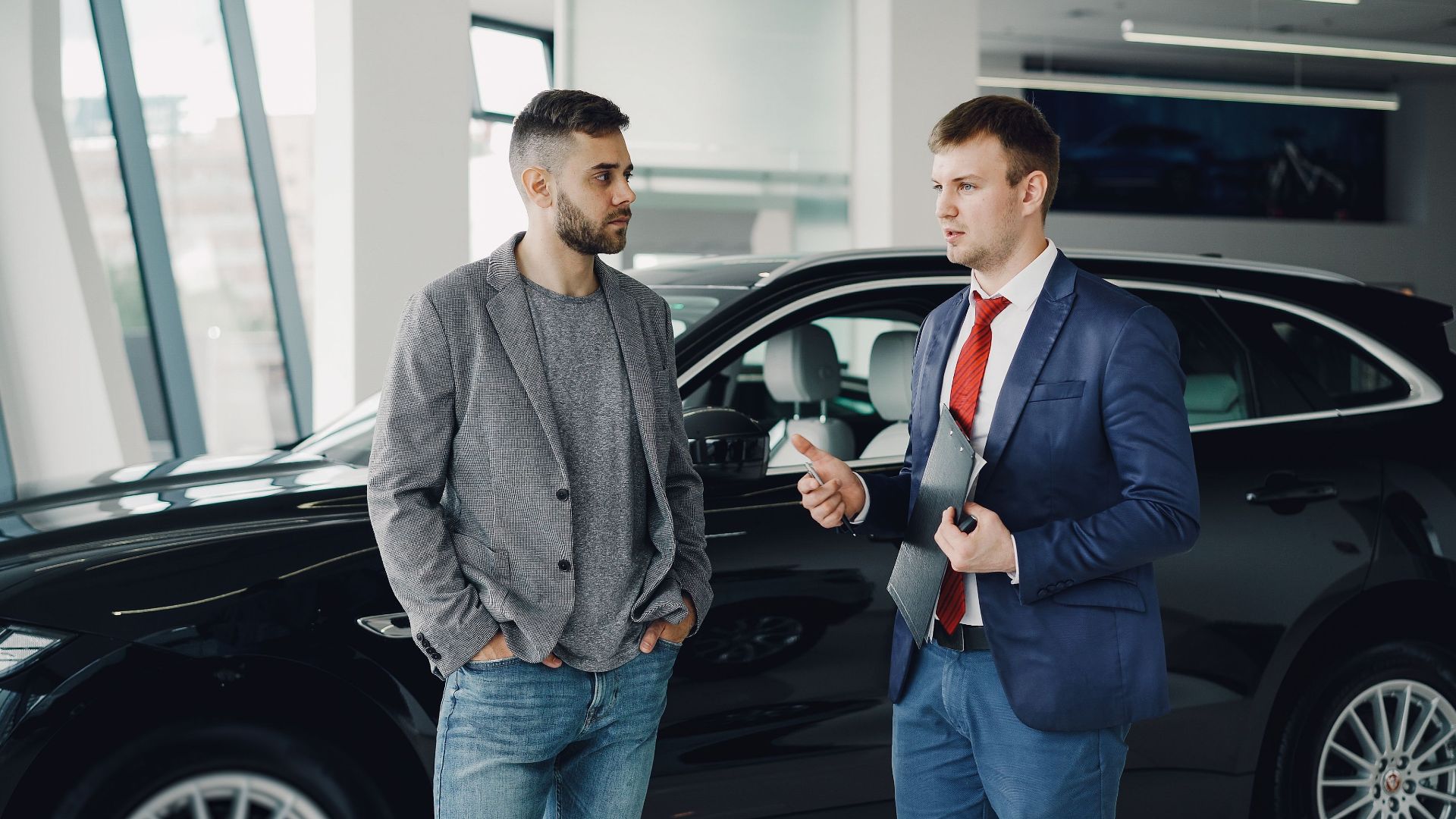 Two men discussing business in a car dealership, standing near a vehicle.