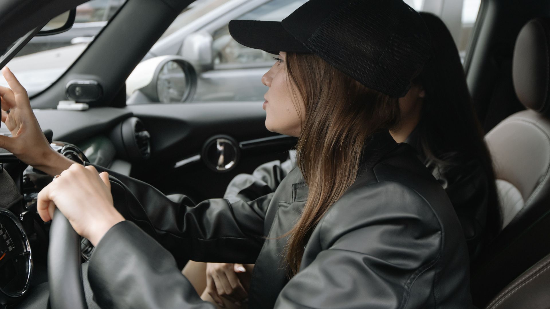 A woman in a leather jacket and cap is driving a modern car interior.