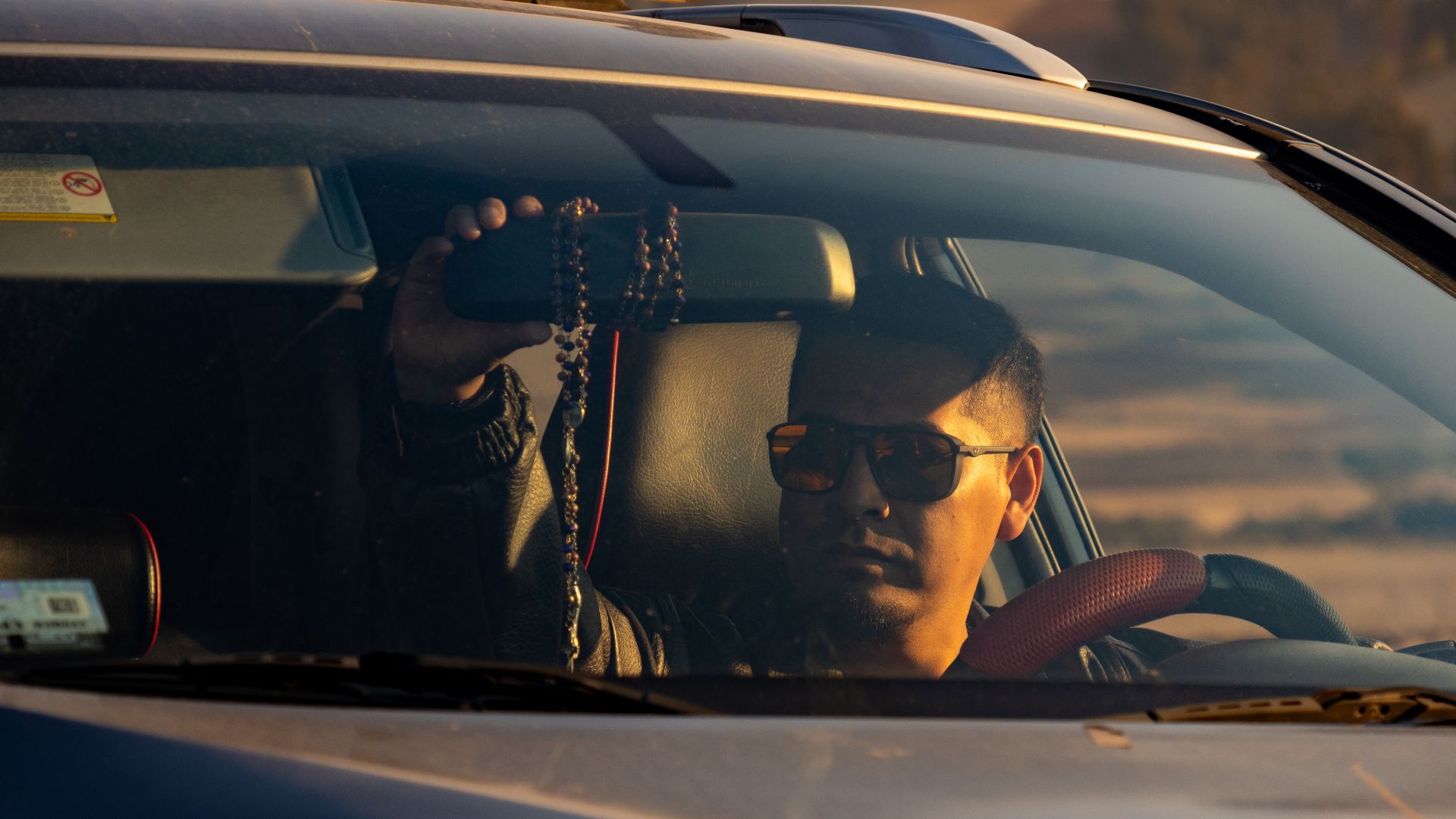 A man wearing sunglasses is seen driving a car at sunset, with a scenic background.