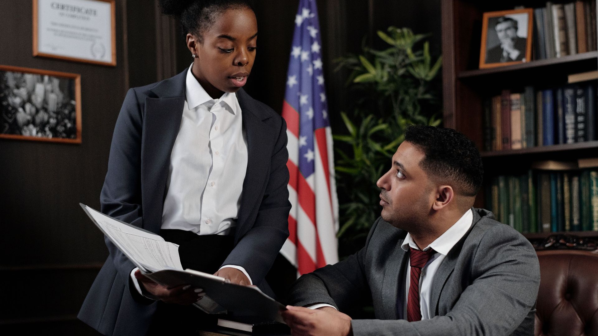 Two professionals discussing documents in an office with an American flag in the background.