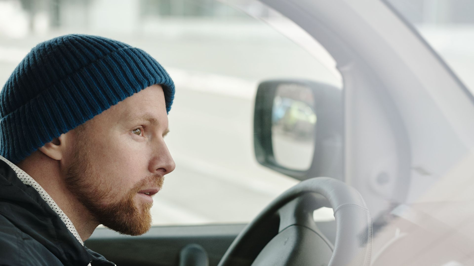 Caucasian man in a knitted cap using a smartphone while sitting in a parked car.