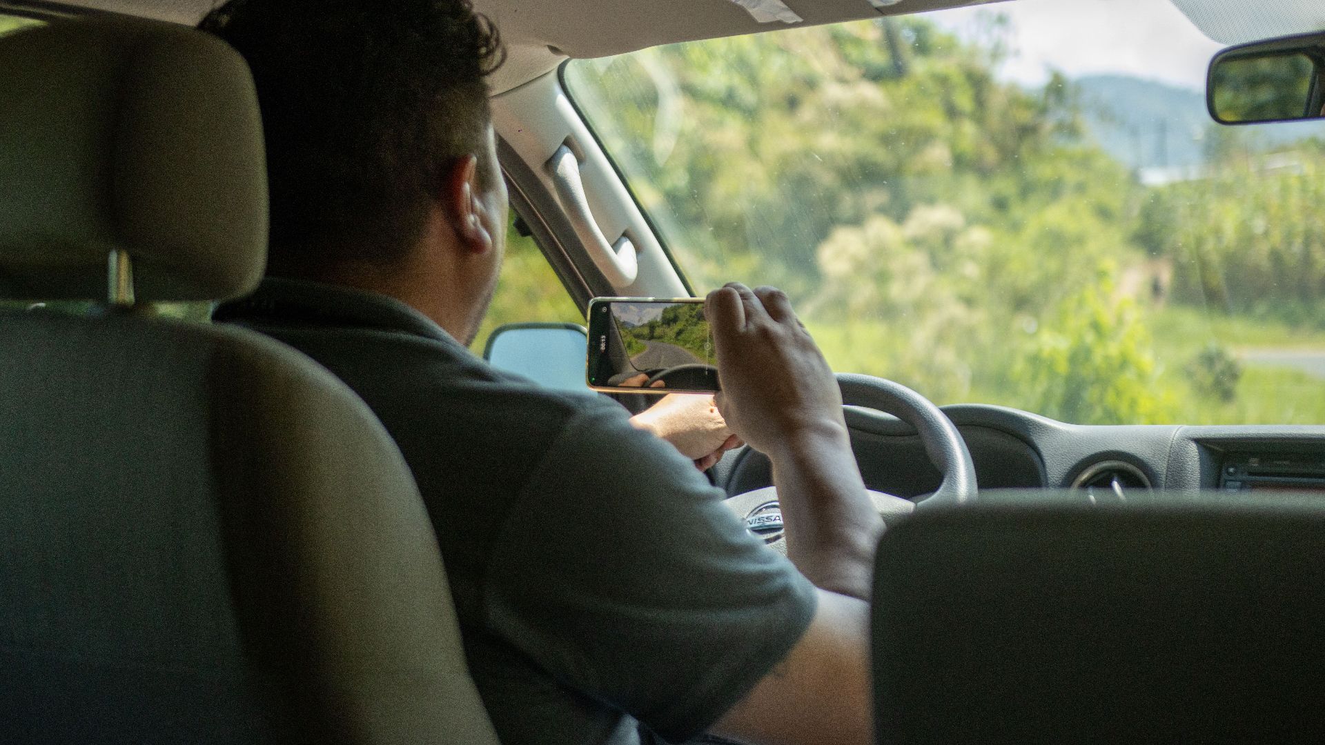 A man uses a smartphone to capture a scene outside while driving in a car interior during the day.