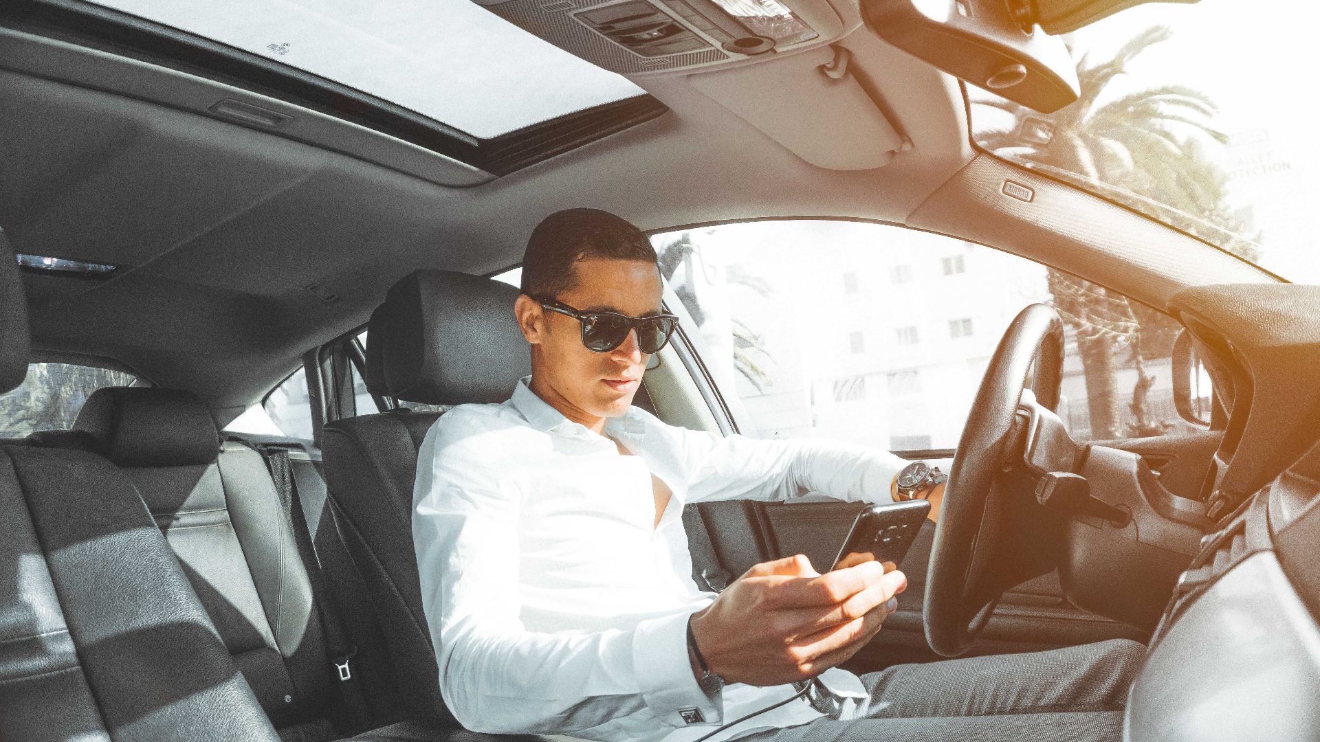 A man wearing sunglasses texts on his phone while driving a car in Morocco.