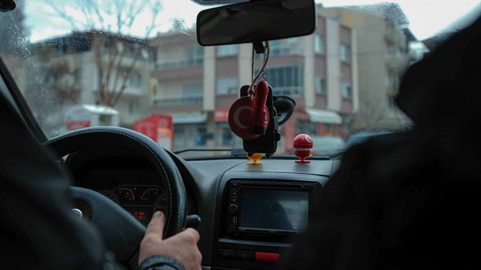 A driver's perspective of a car interior navigating through city streets with focus on the dashboard and rearview mirror.