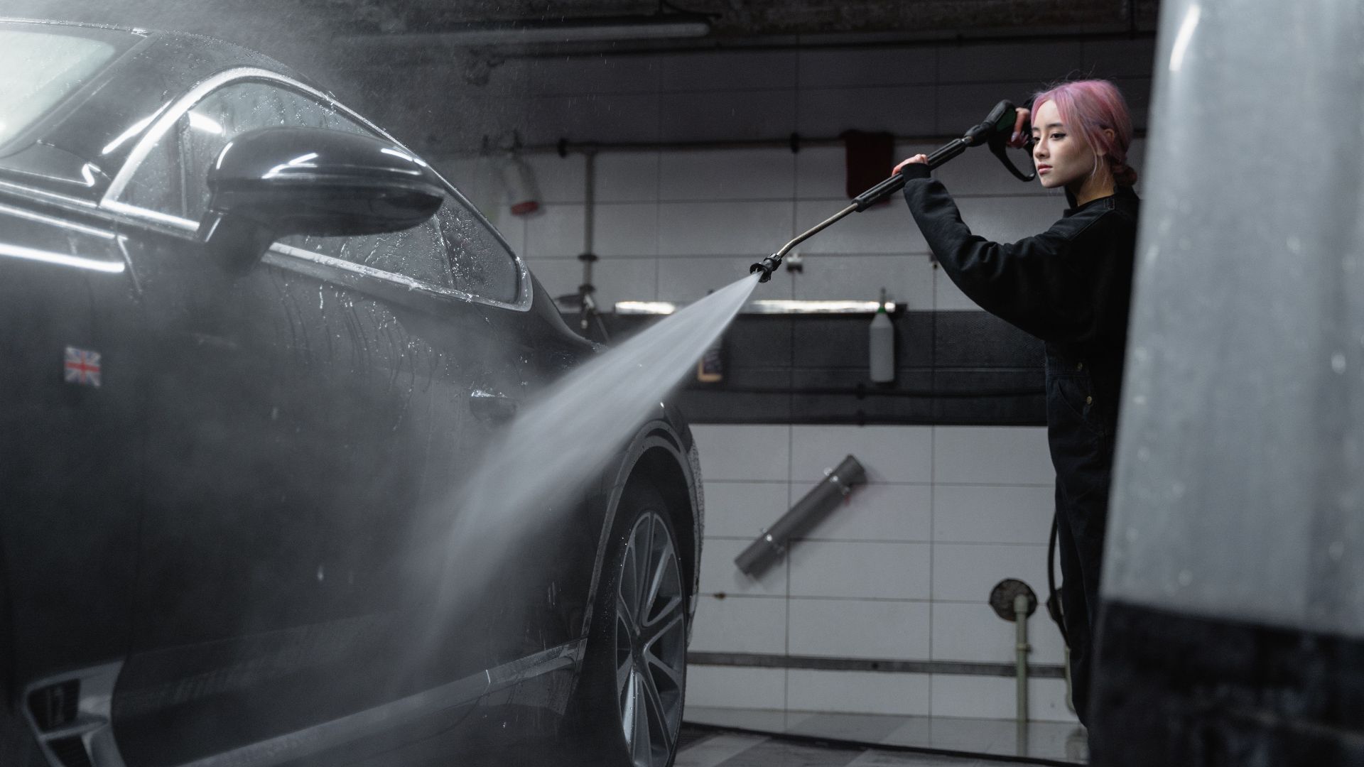 Woman power washing a black car in an indoor garage using a high-pressure washer.