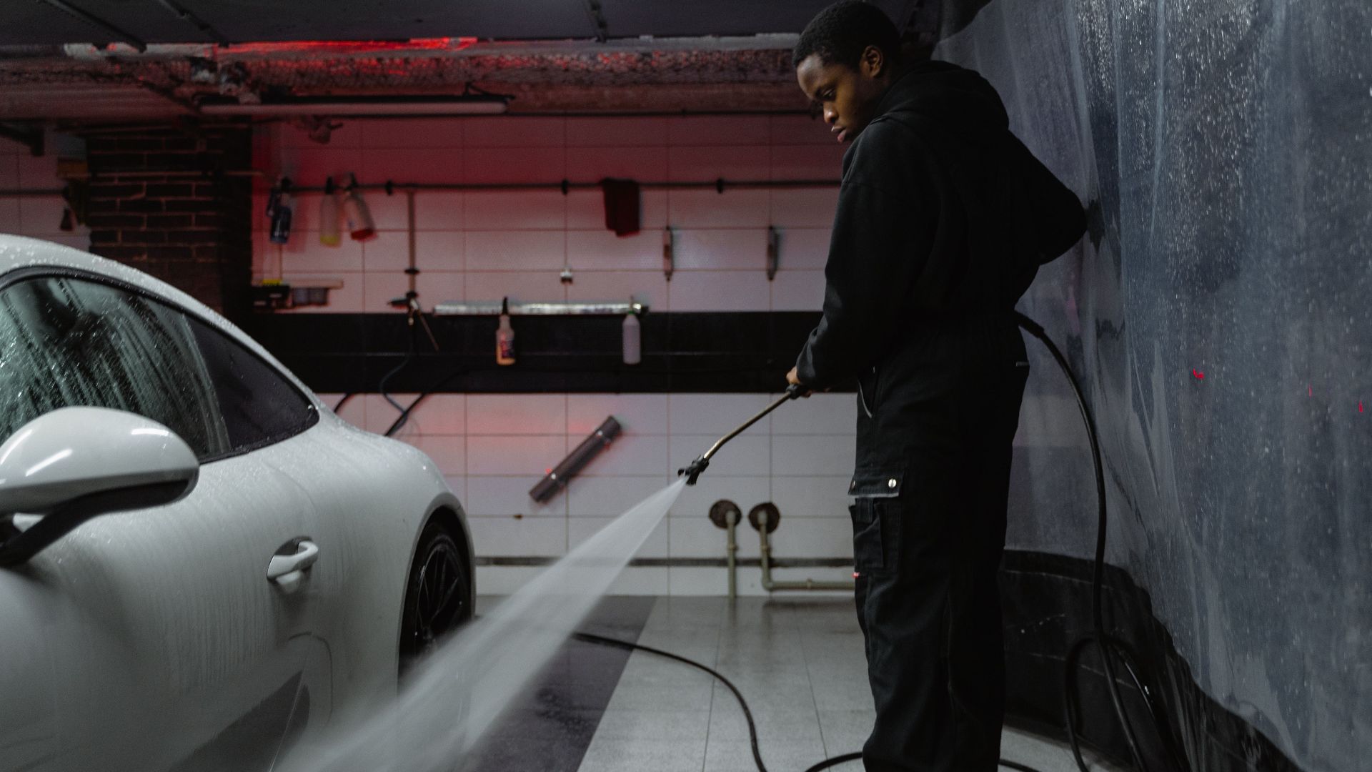 African American man cleaning a white car with a pressure hose in an indoor garage setting.