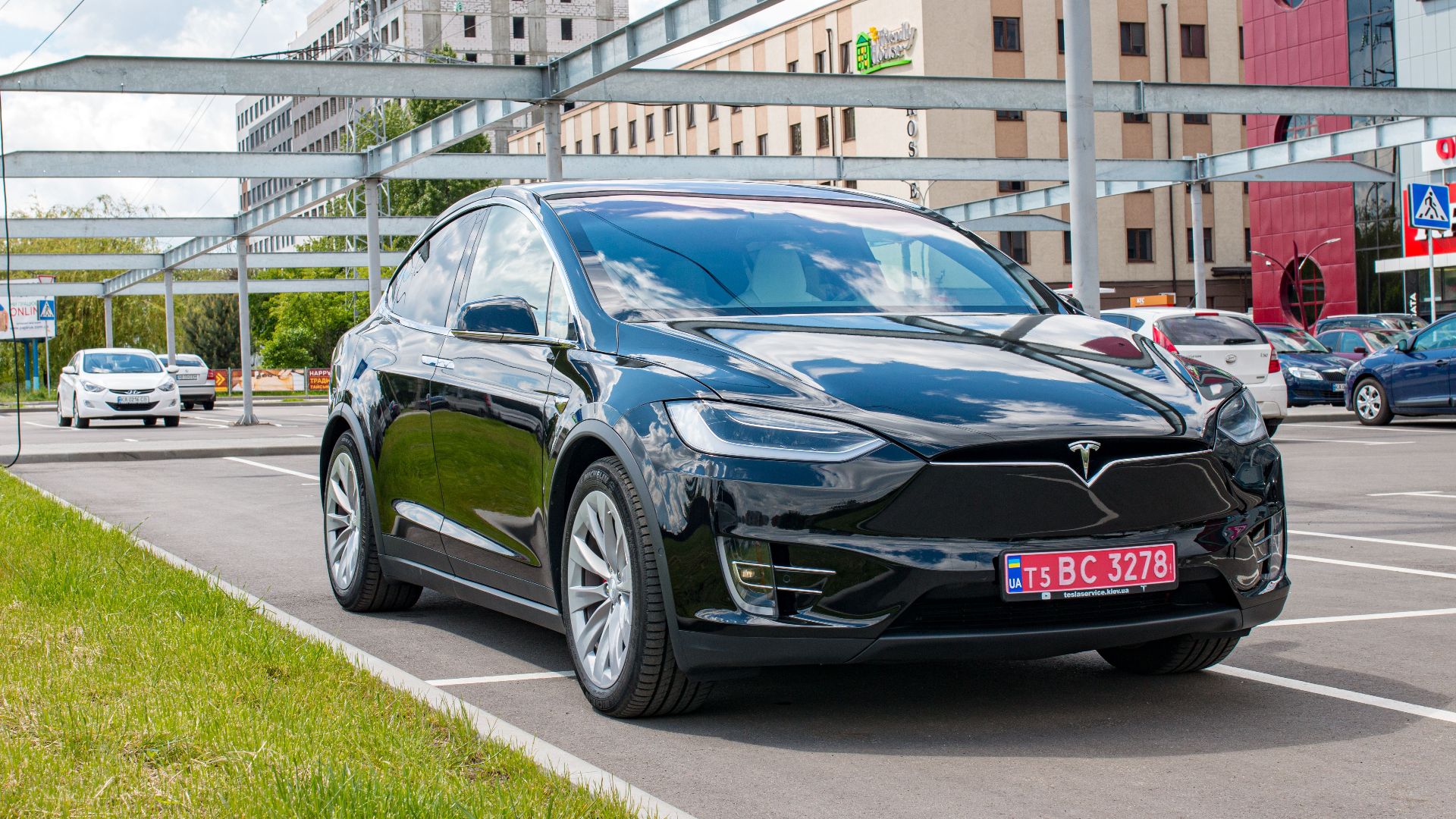 Black Tesla Model X parked in a city lot reflecting buildings with a cloudy sky.