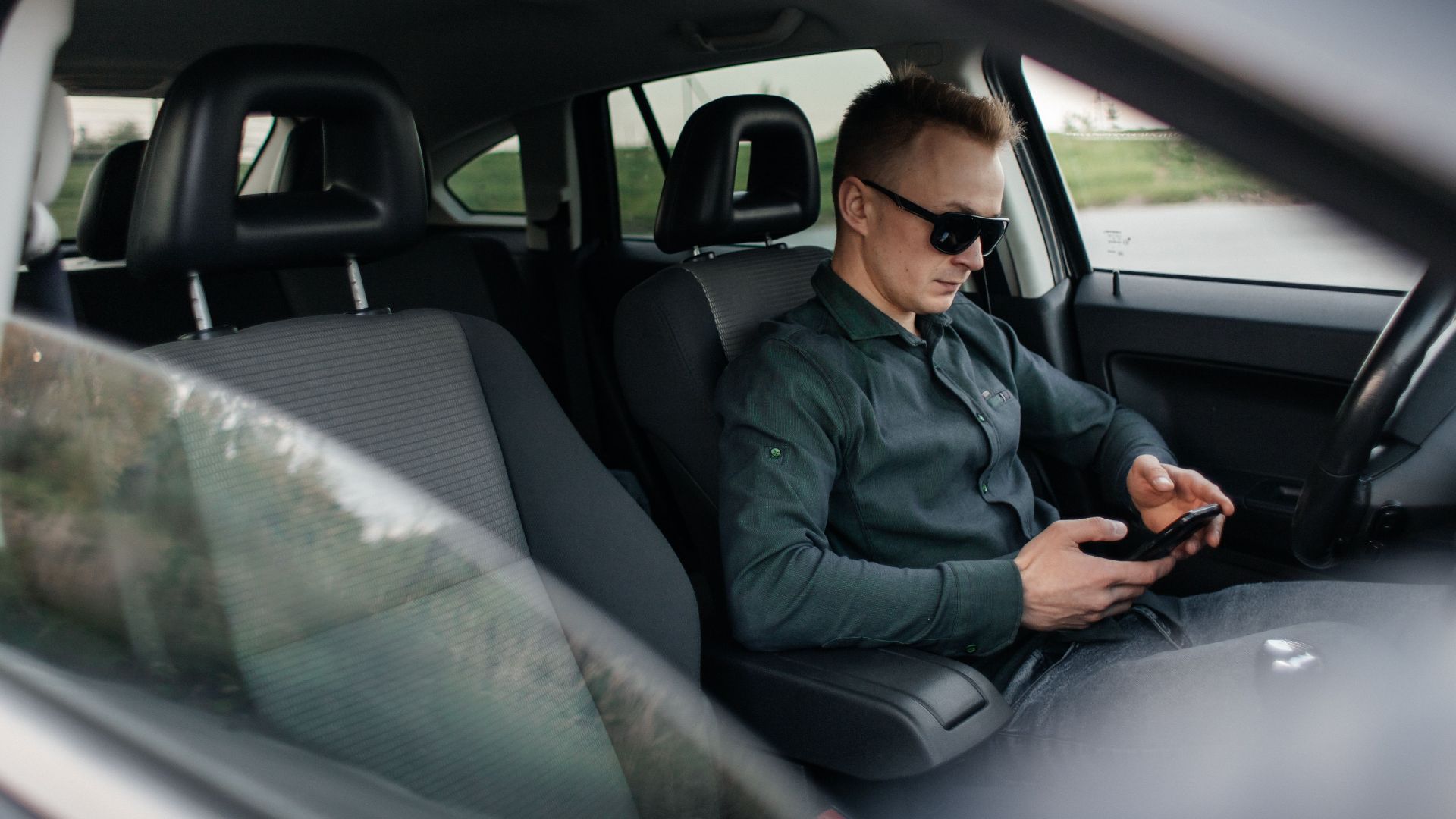 Young man using smartphone inside a parked car, wearing sunglasses.