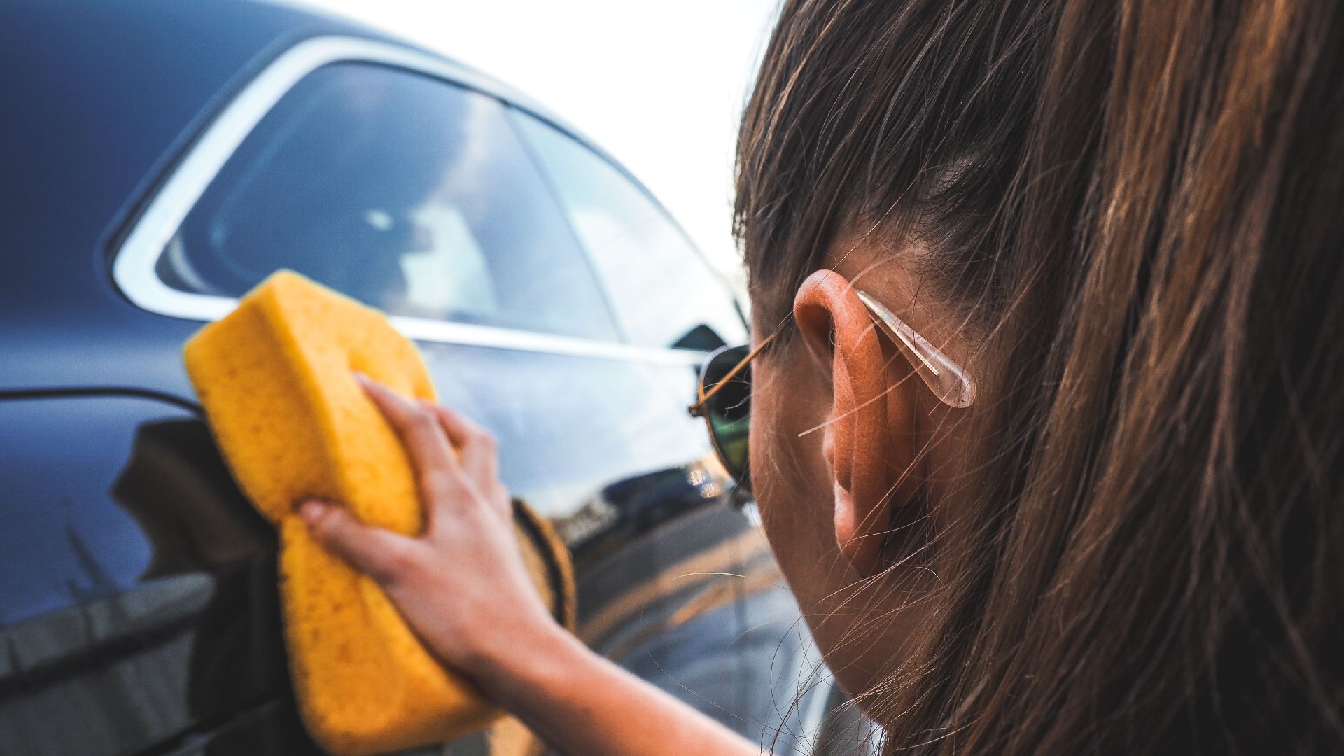 Close-up of a woman handwashing a black car using a yellow sponge, highlighting cleaning process.