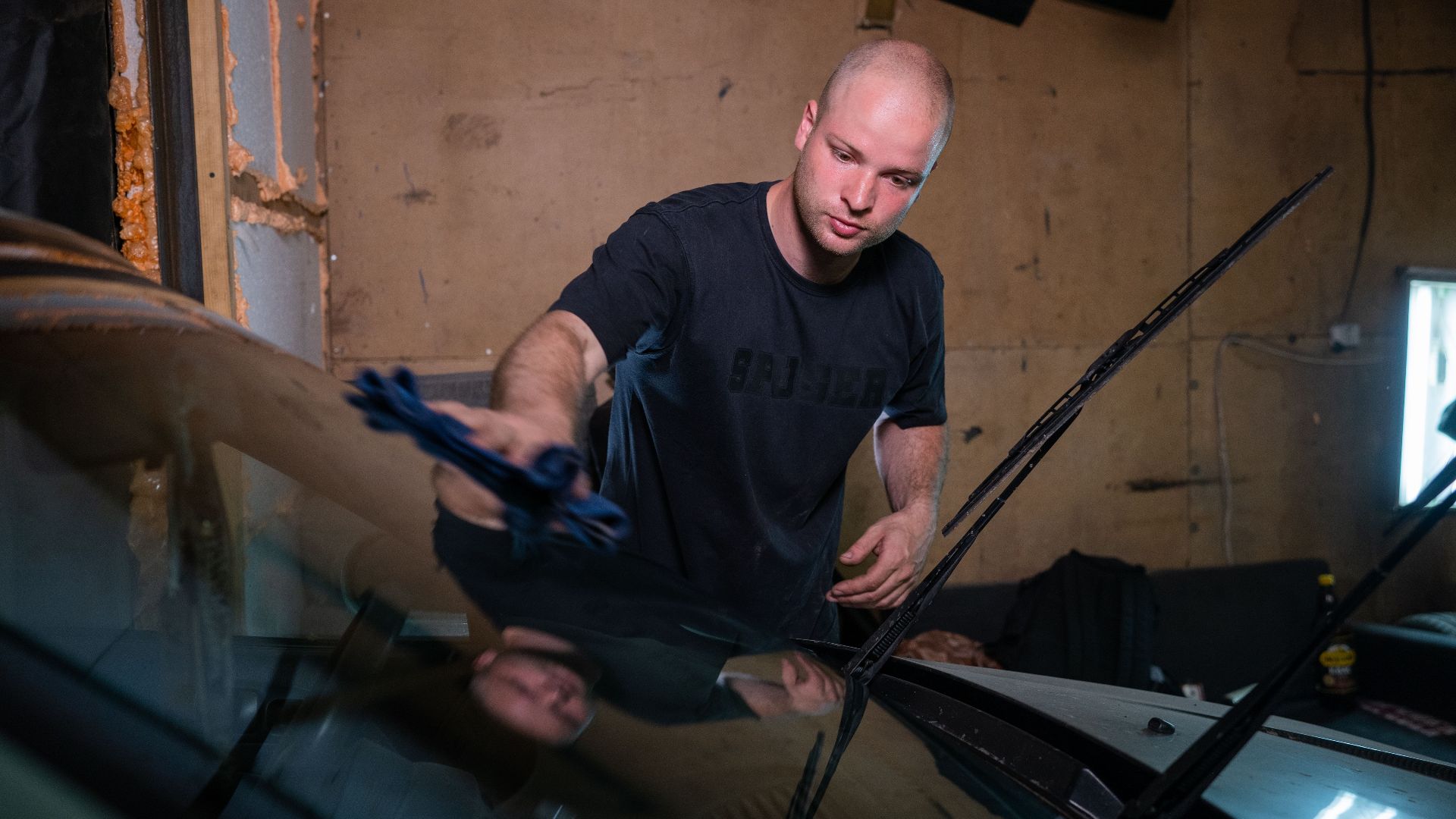 A man wipes a car windshield inside a garage, focused on cleaning.