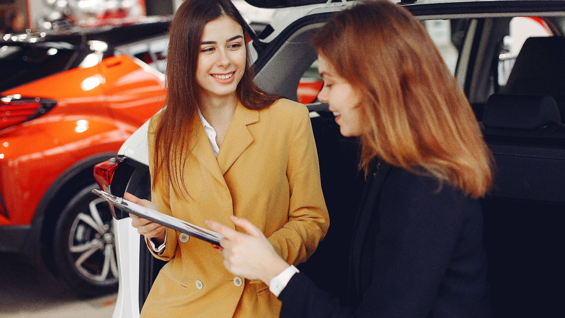 Positive smiling agent in trendy formal light brown suit discussing with happy excited female customer details of contract while leaning on car in showroom