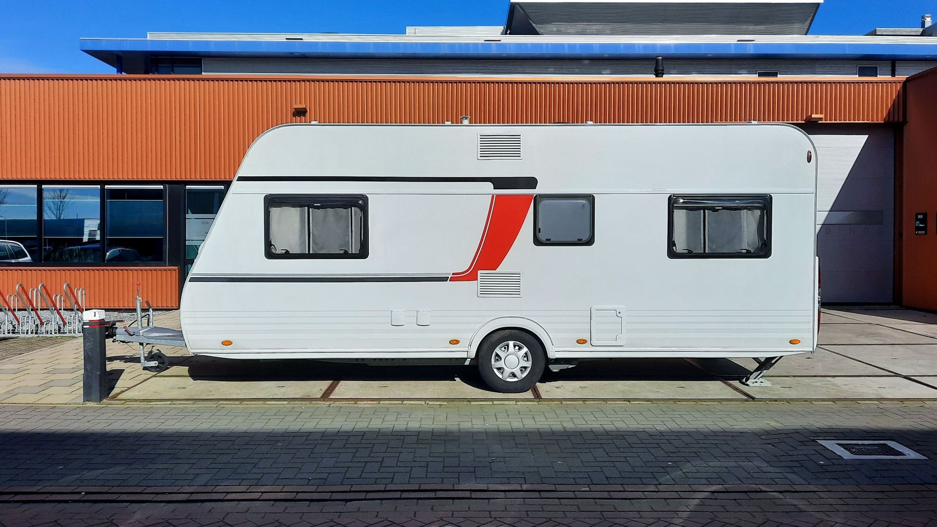 A sleek white caravan with red accents parked next to an industrial building under a clear blue sky.