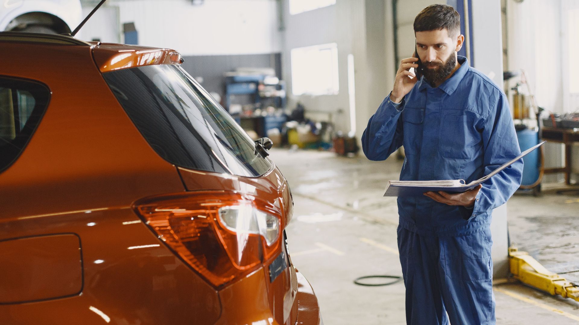Mechanic in a blue uniform talks on phone beside a red car in an auto repair shop.